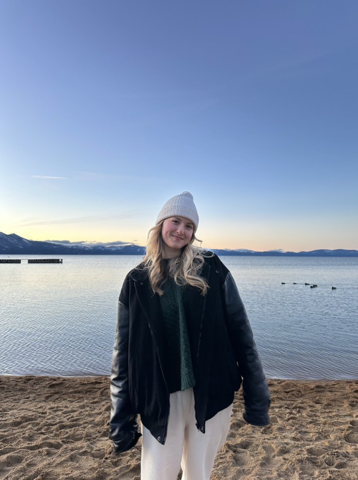 A young woman with blond hair, wearing a gray beanie, black jacket, green sweater, and white pants, standing on a sandy beach by a lake during sunset with mountains in the background.