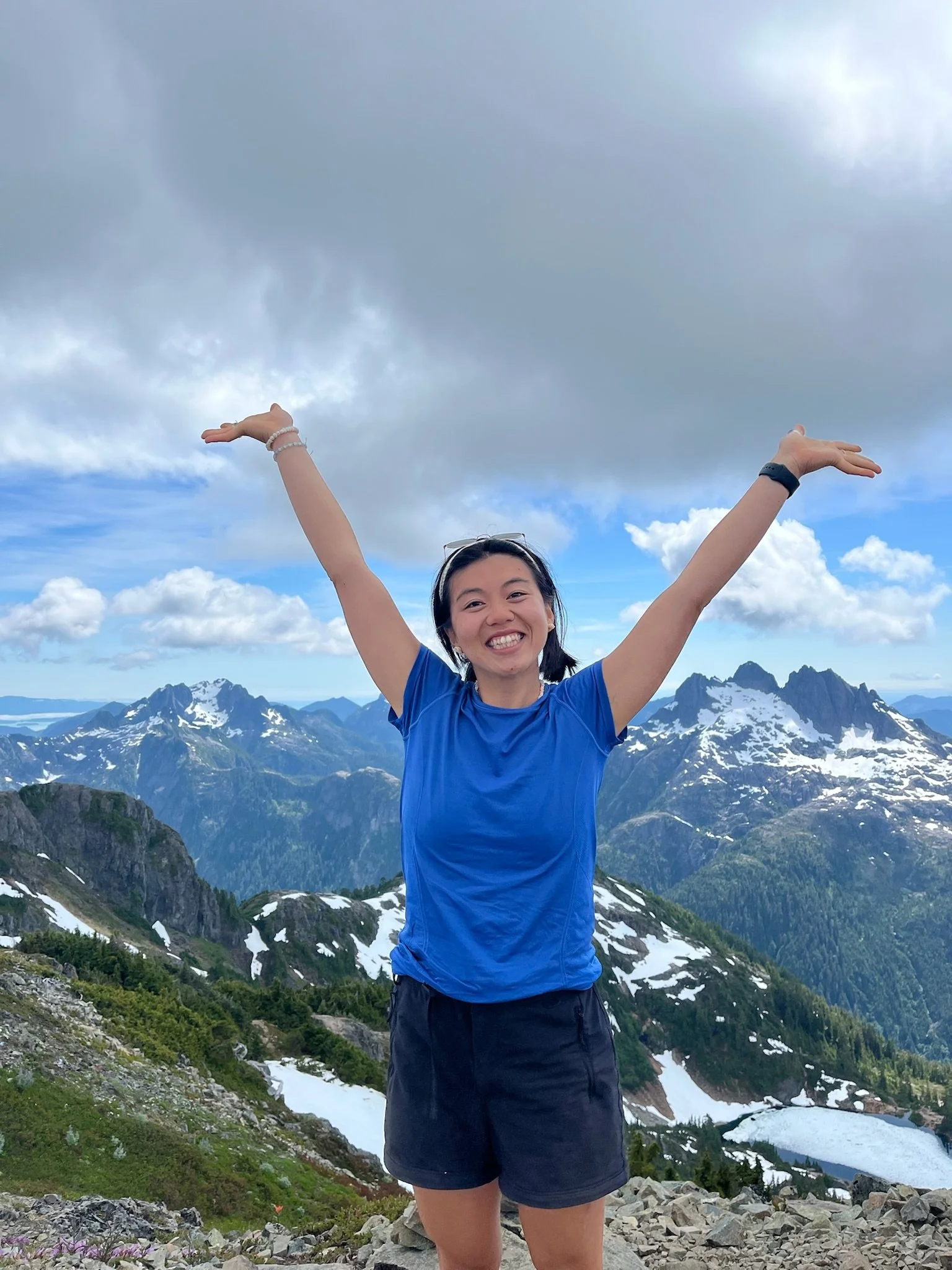 Smiling woman with arms raised in front of a mountain range with snow patches, cloudy sky, and some blue sky visible.