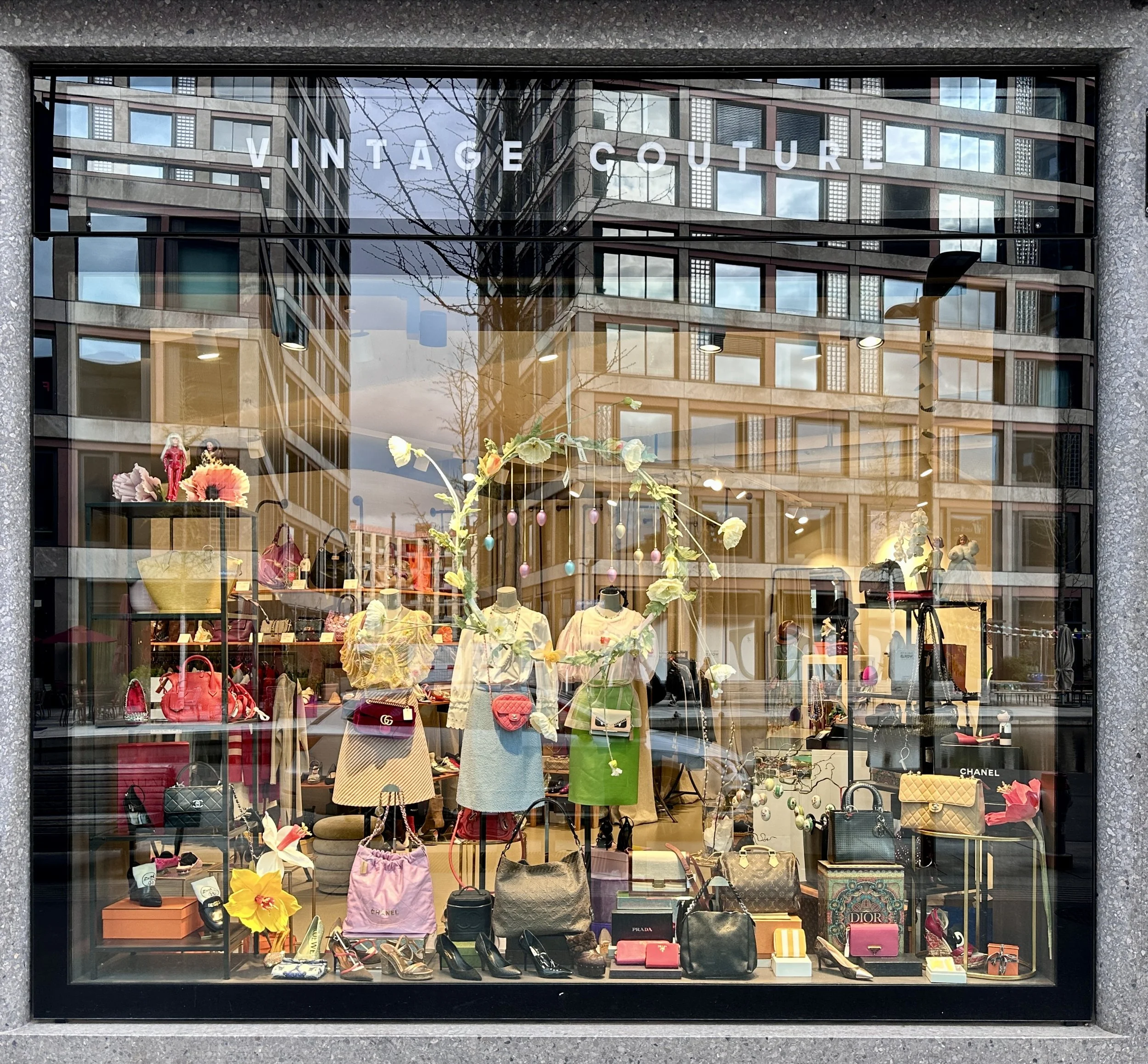 Schaufenster eines Modegeschäfts mit Vintage Couture, zeigt Kleidung, Taschen, Schuhe und Accessoires, reflektierende Fenster mit Stadtansicht, inklusive Hochhäuser und Bäume.