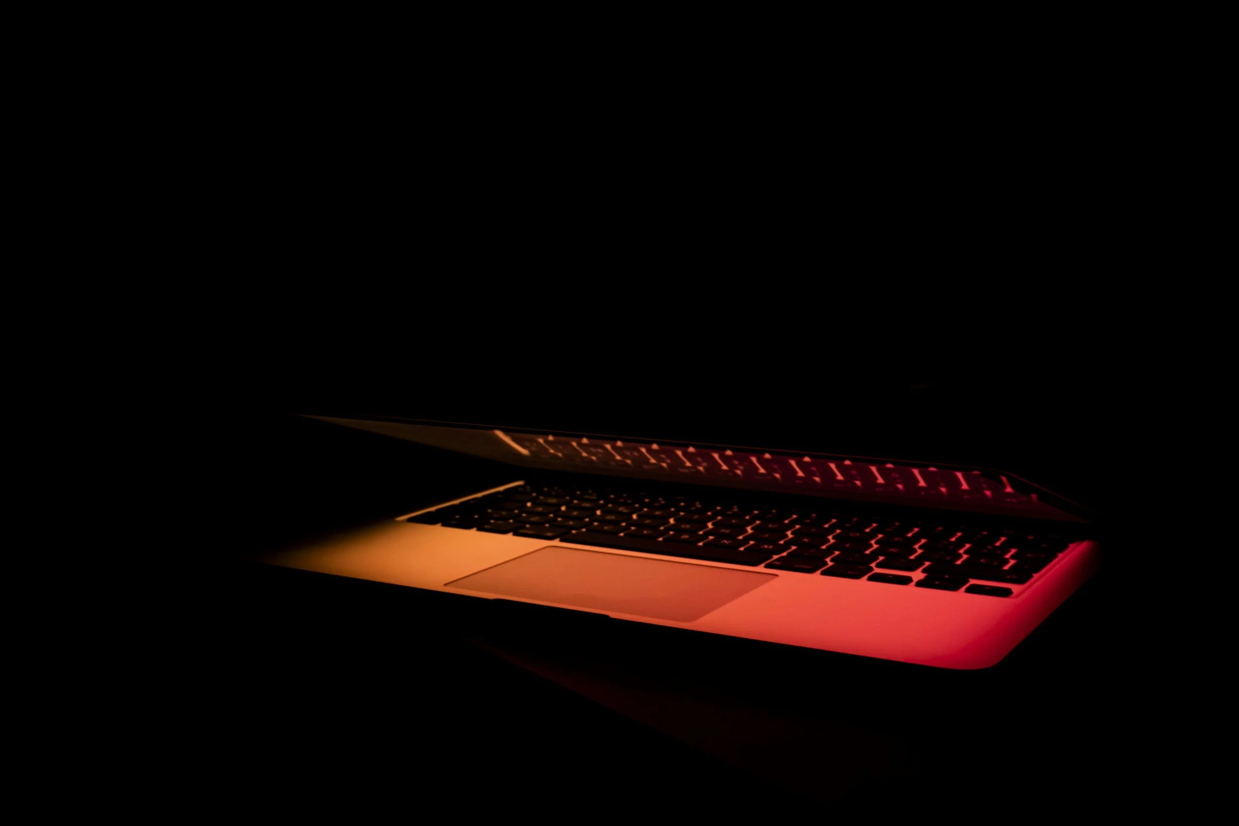A laptop computer with a glowing keyboard illuminated in red and orange light, partially open in a dark environment.