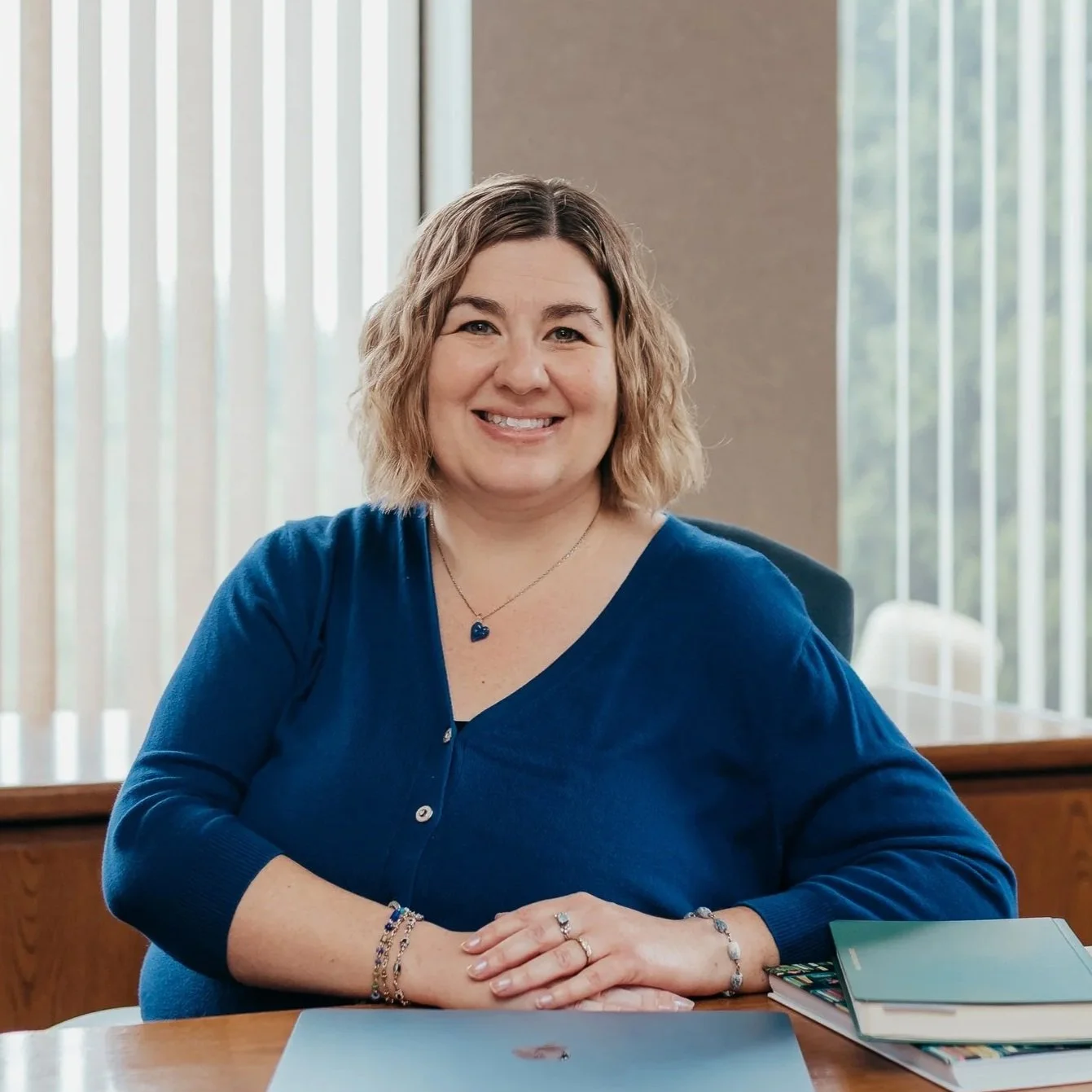 A woman with shoulder-length blonde curly hair sitting at a desk with a smile, wearing a blue top and jewelry, in an office with large windows and vertical blinds in the background.