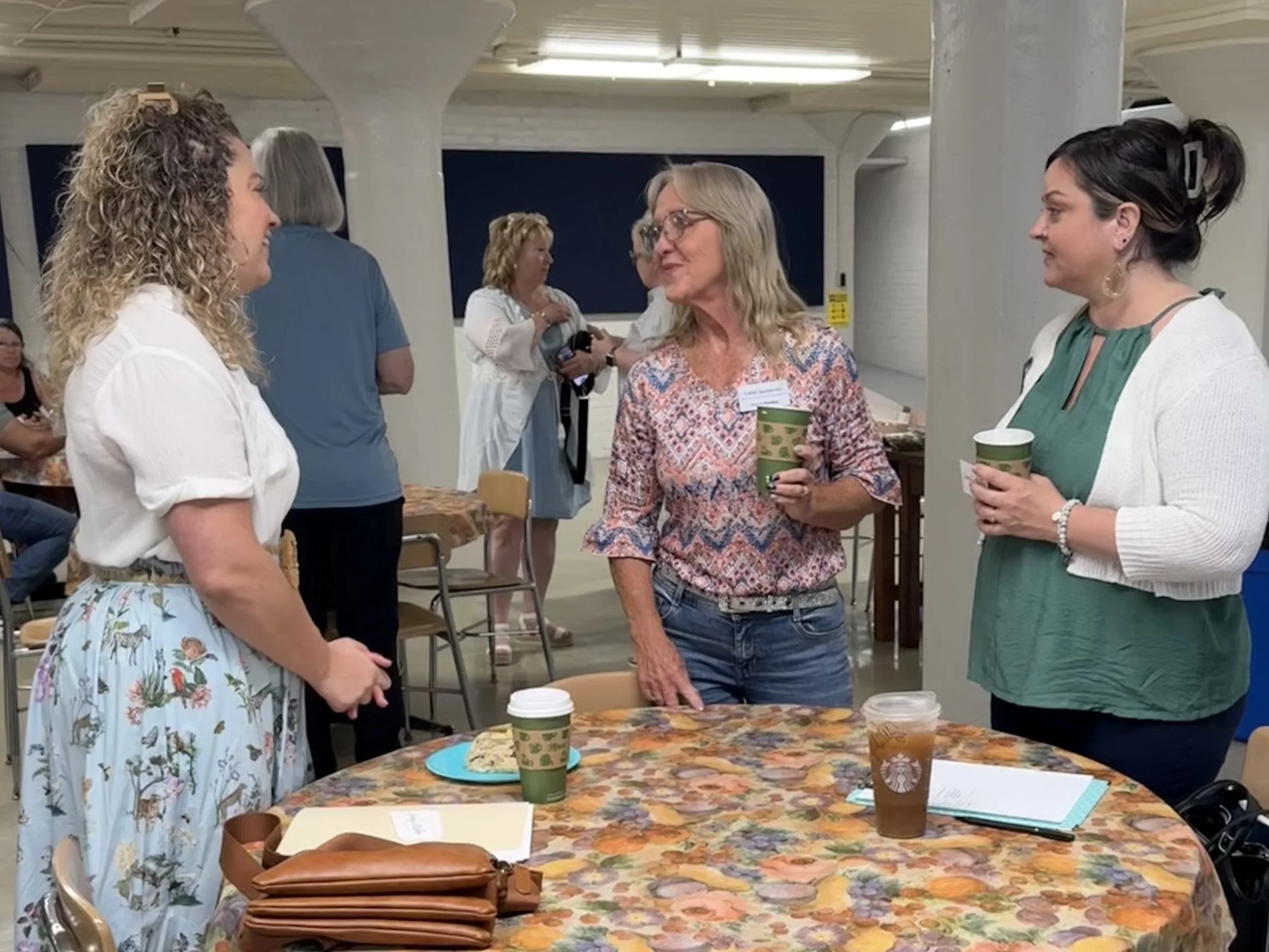 People in conversation at a social gathering in a room with a long table covered with a floral tablecloth, coffee cups, and snacks. The atmosphere appears friendly and relaxed.