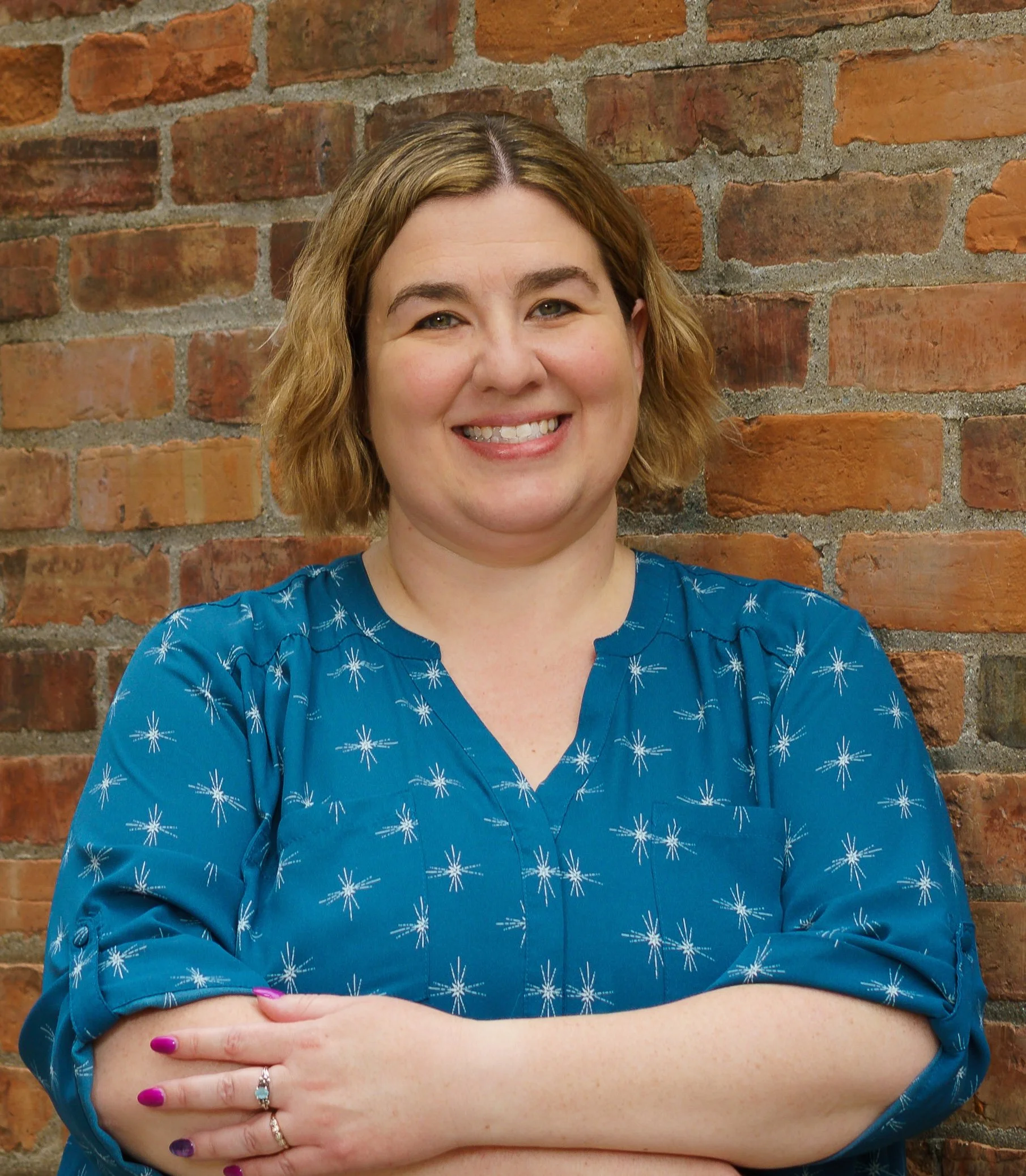 A woman with shoulder-length light brown hair, smiling, standing in front of a brick wall. She is wearing a blue blouse with white starburst patterns, with her arms crossed, showing pink and purple nail polish and rings on her fingers.