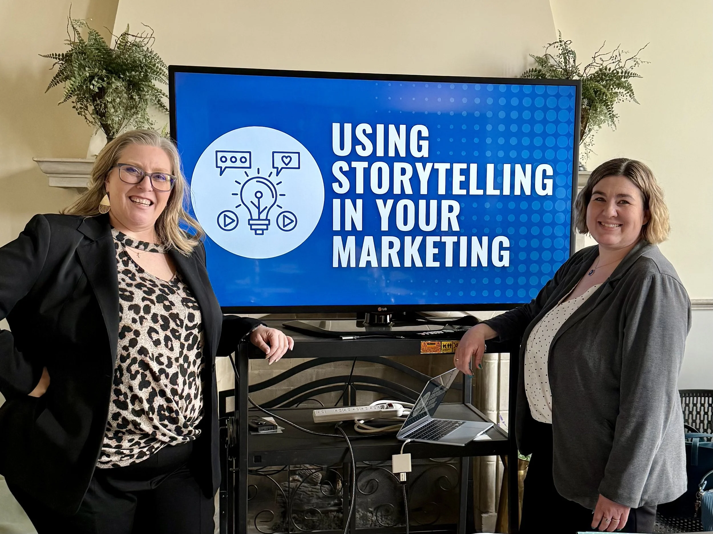Two women standing in front of a large screen that displays the text "Using Storytelling in Your Marketing" along with a graphic of a light bulb, speech bubbles, and play buttons. One woman has blonde hair, glasses, and is wearing a black blazer with a leopard print top. The other woman has brown hair and is wearing a gray blazer over a polka-dotted top.