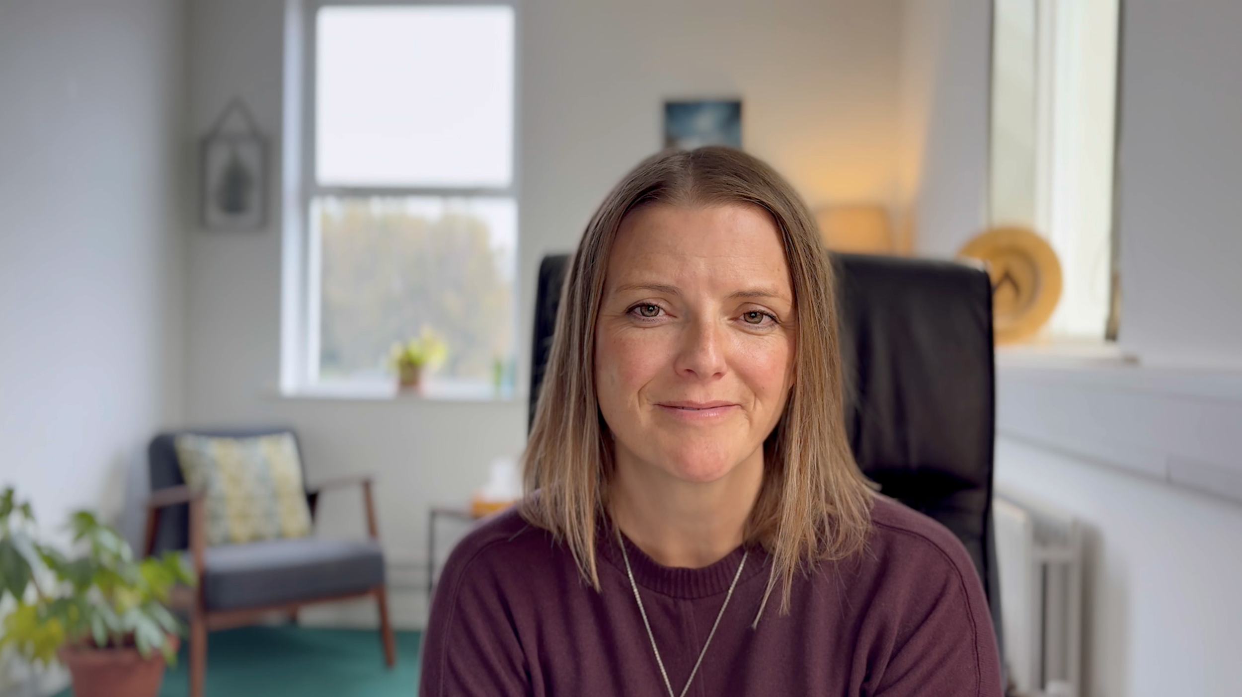A professional female sitting in a therapy room with soft lighting and consultation chairs slight out of focus behind her.