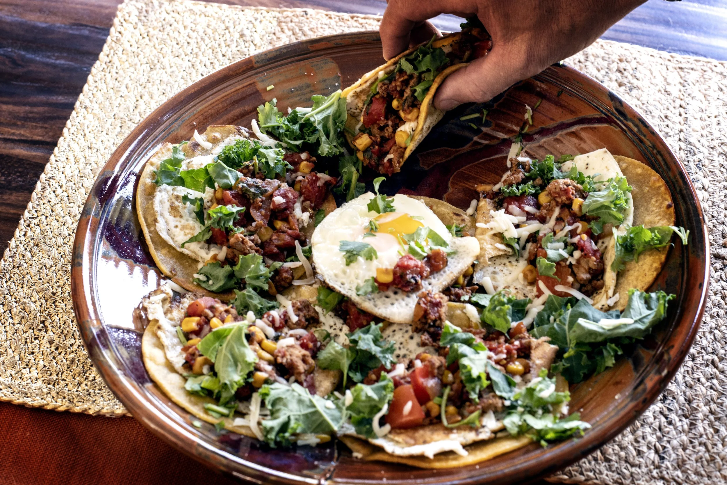 hand grabbing a taco from a plate of shakshuka tacos with corn tortillas, fried eggs, kale, tomatoes, red bell peppers, and ground pheasant sausage