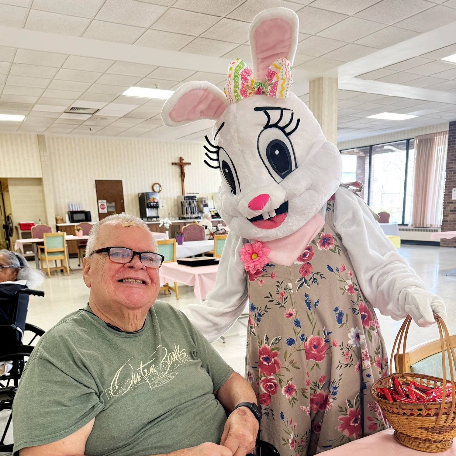 A man smiling and wearing glasses sitting next to a person in a bunny costume at an indoor event with tables, chairs, and a cross on the wall in the background.