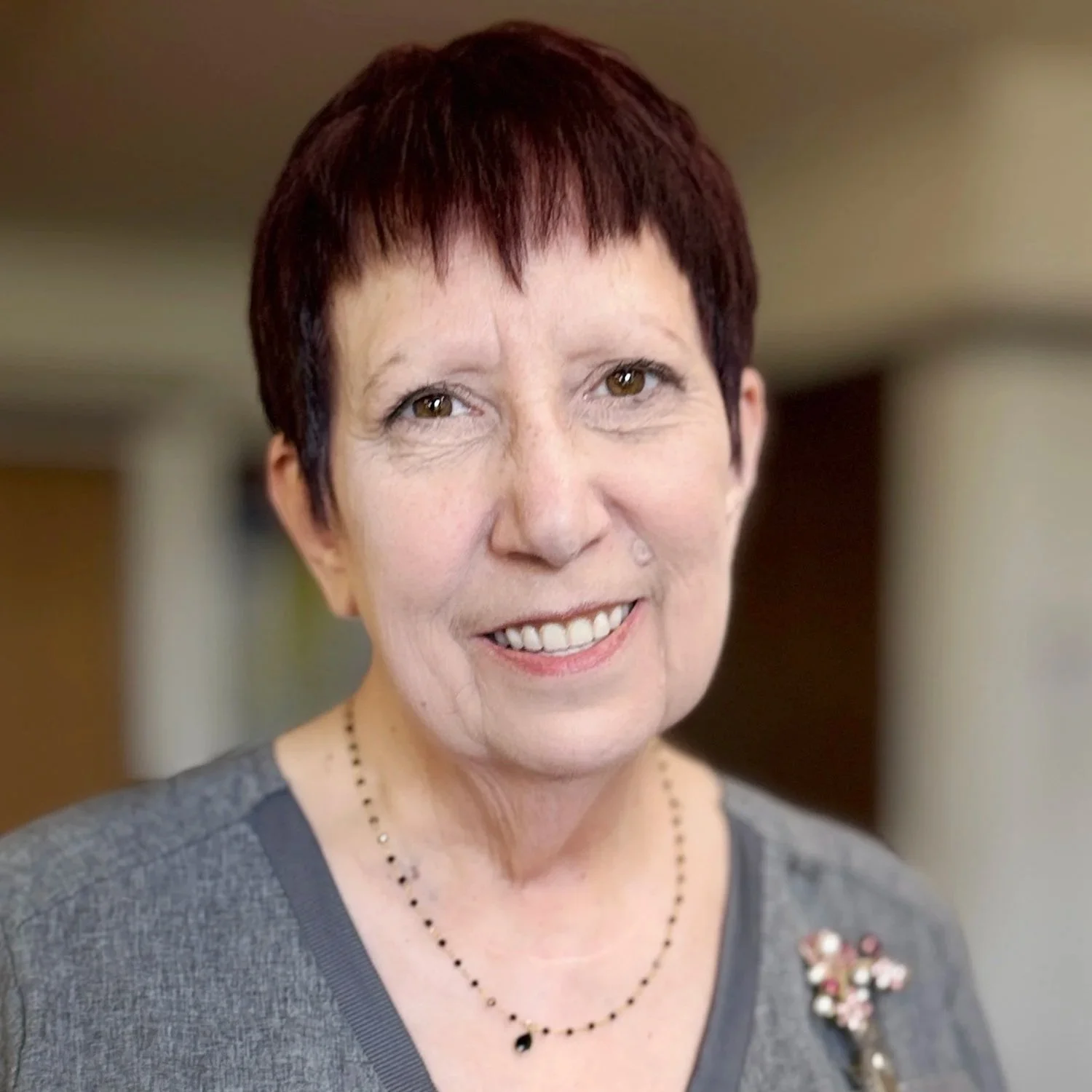 A smiling middle-aged woman with short dark red hair, wearing a gray top, a necklace, and a brooch shaped like a flower.
