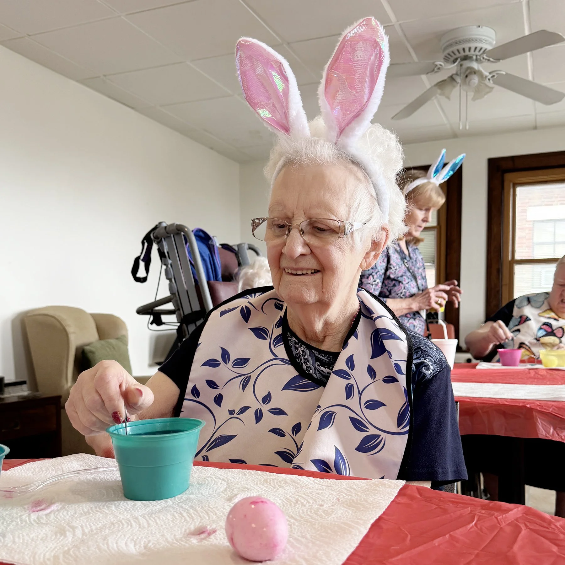 An elderly woman wearing bunny ears made of pink and white fabric, smiling as she dyes Easter eggs at a table with a cup of dye and a white paper towel. Other women are visible in the background, participating in the activity in a room with beige walls, wooden trim, and windows.