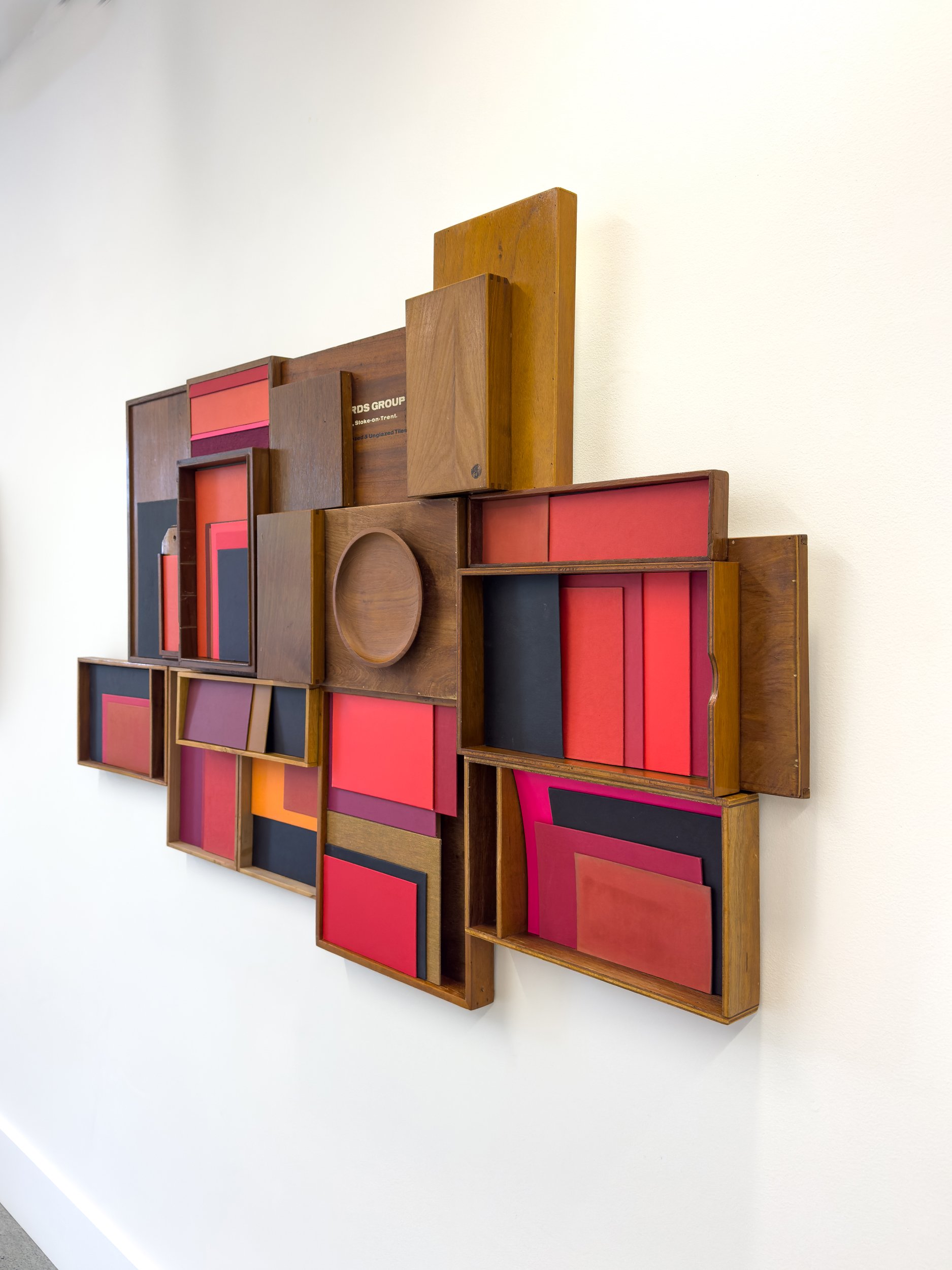 Wooden wall sculpture with red, pink, and black rectangular panels arranged in a geometric pattern. Minimalist and modern design on a white wall.