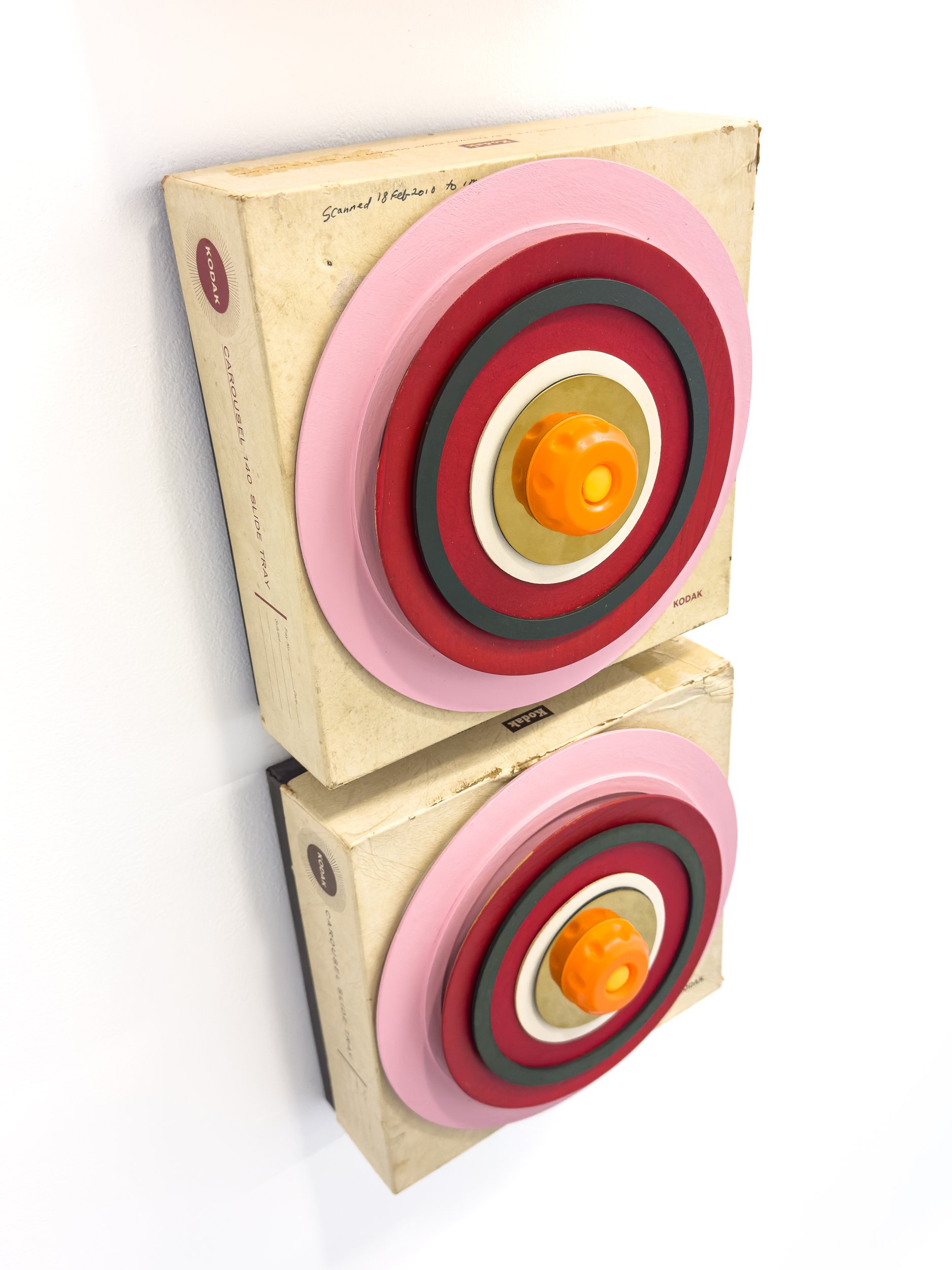 Two stacked wooden targets, each with concentric circles in pink, red, and black, featuring an orange knob at the center, on a textured beige background.
