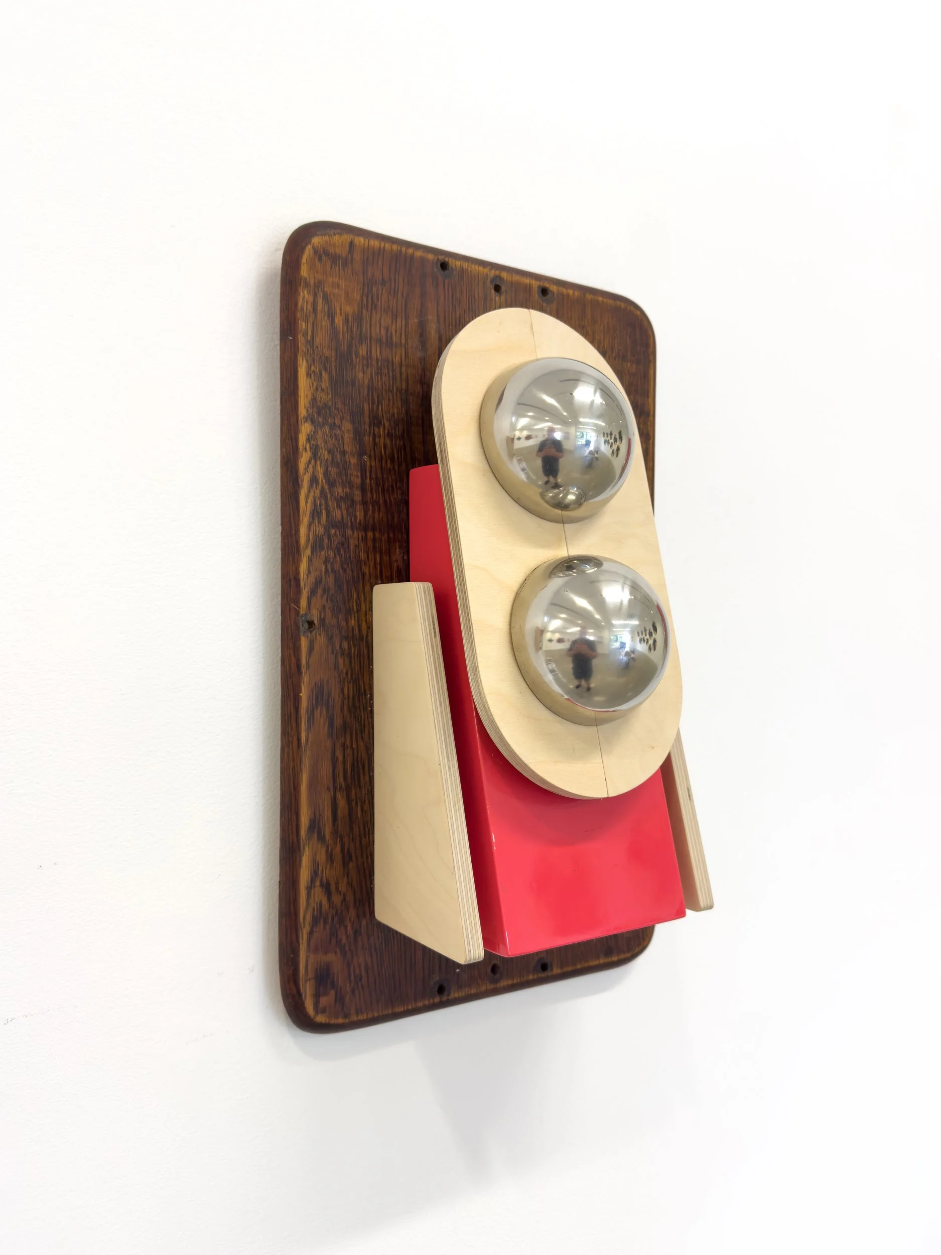 Wooden art piece resembling a robot face; two metallic spheres as eyes, mounted on a wooden plaque with red and beige accents, creating a playful tone.