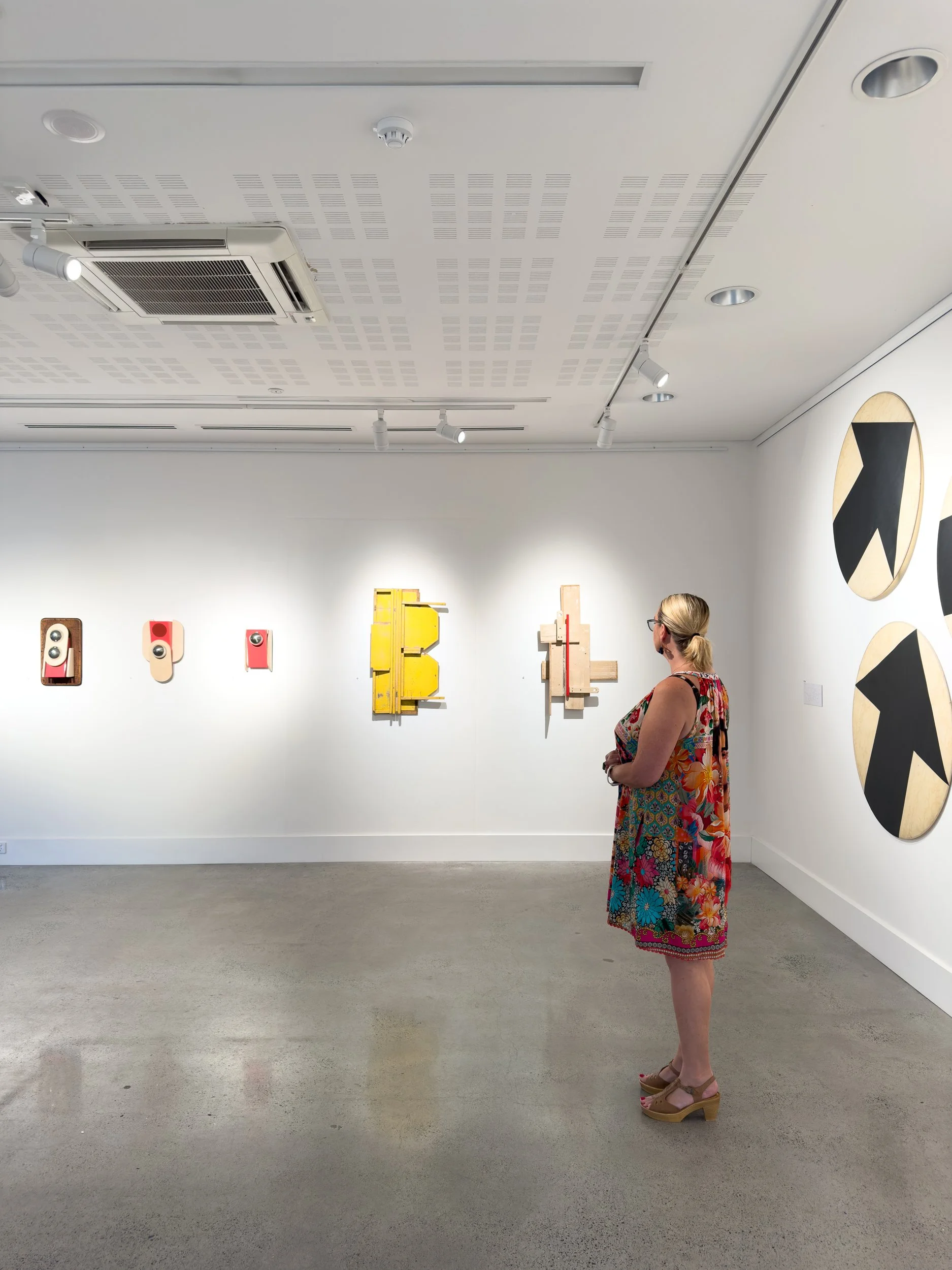 A woman in a colorful dress observes abstract art pieces on a white gallery wall. The artworks feature bold shapes and vibrant colors, creating a contemplative mood.