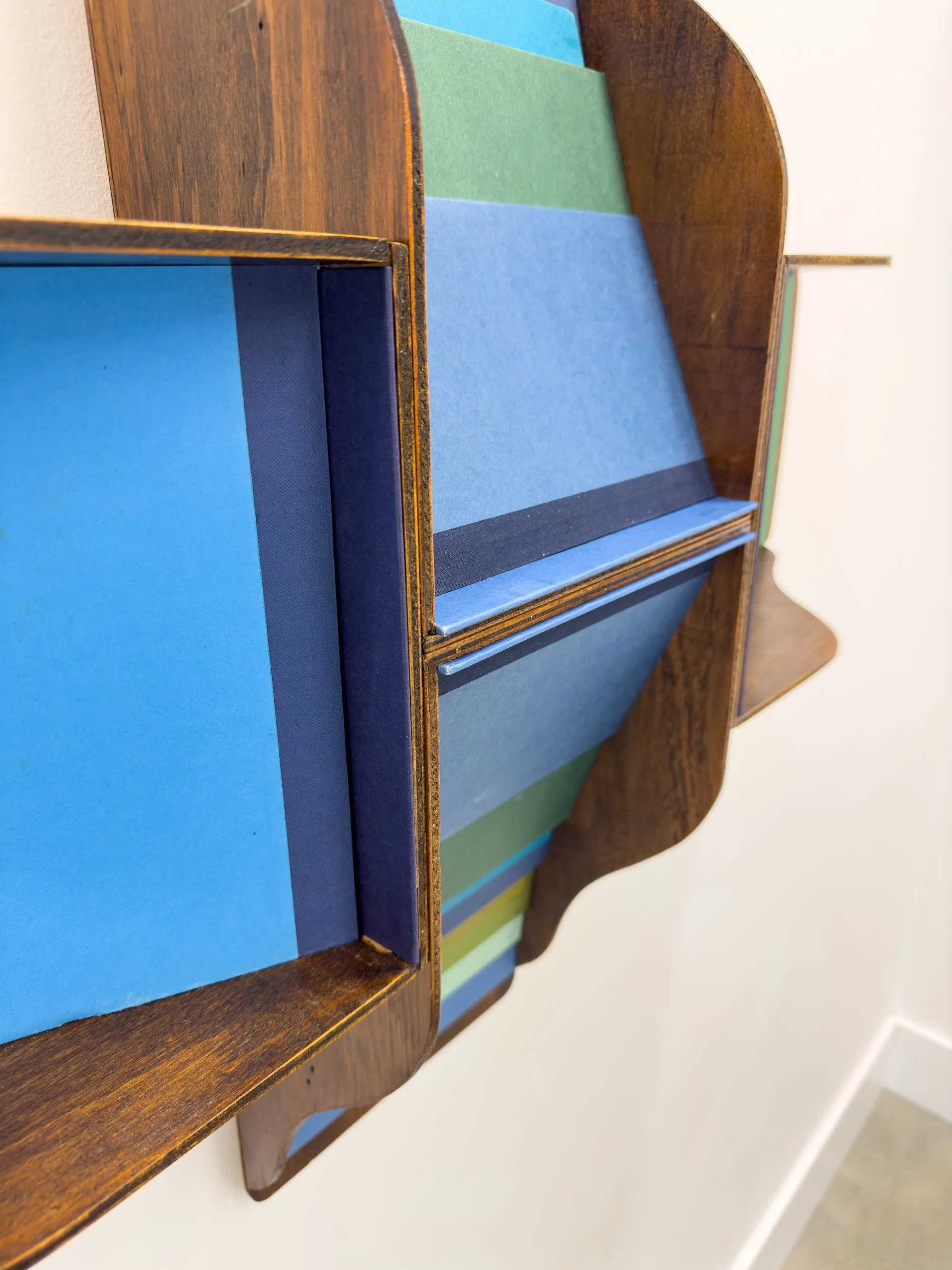 Wall-mounted wooden shelf with colorful striped backing. Blue, green, and teal panels create a vibrant geometric pattern. Modern and artistic design.