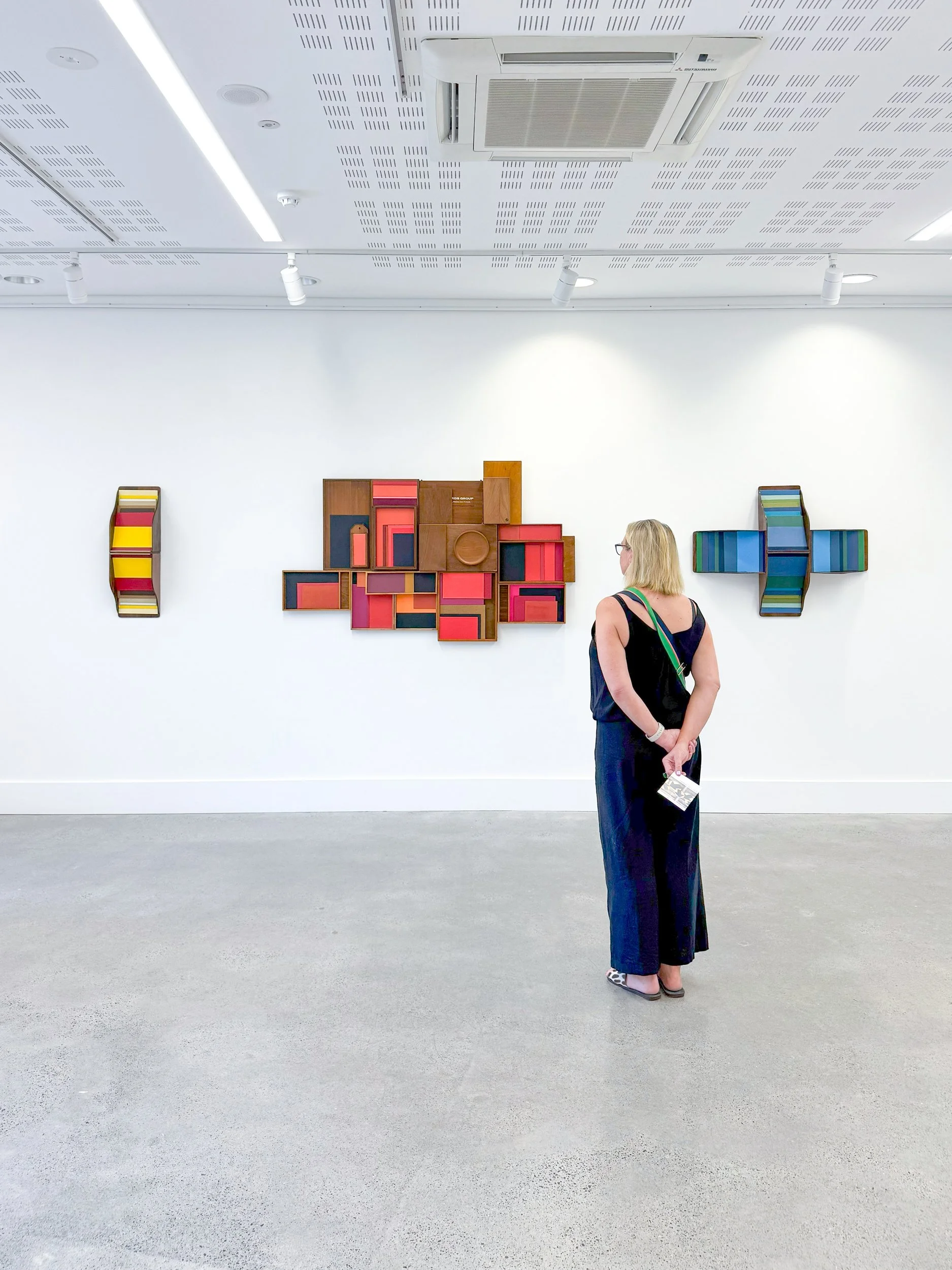 A woman in a black dress observes colorful abstract wall art in a bright gallery. The artworks feature geometric shapes in red, yellow, and blue tones.