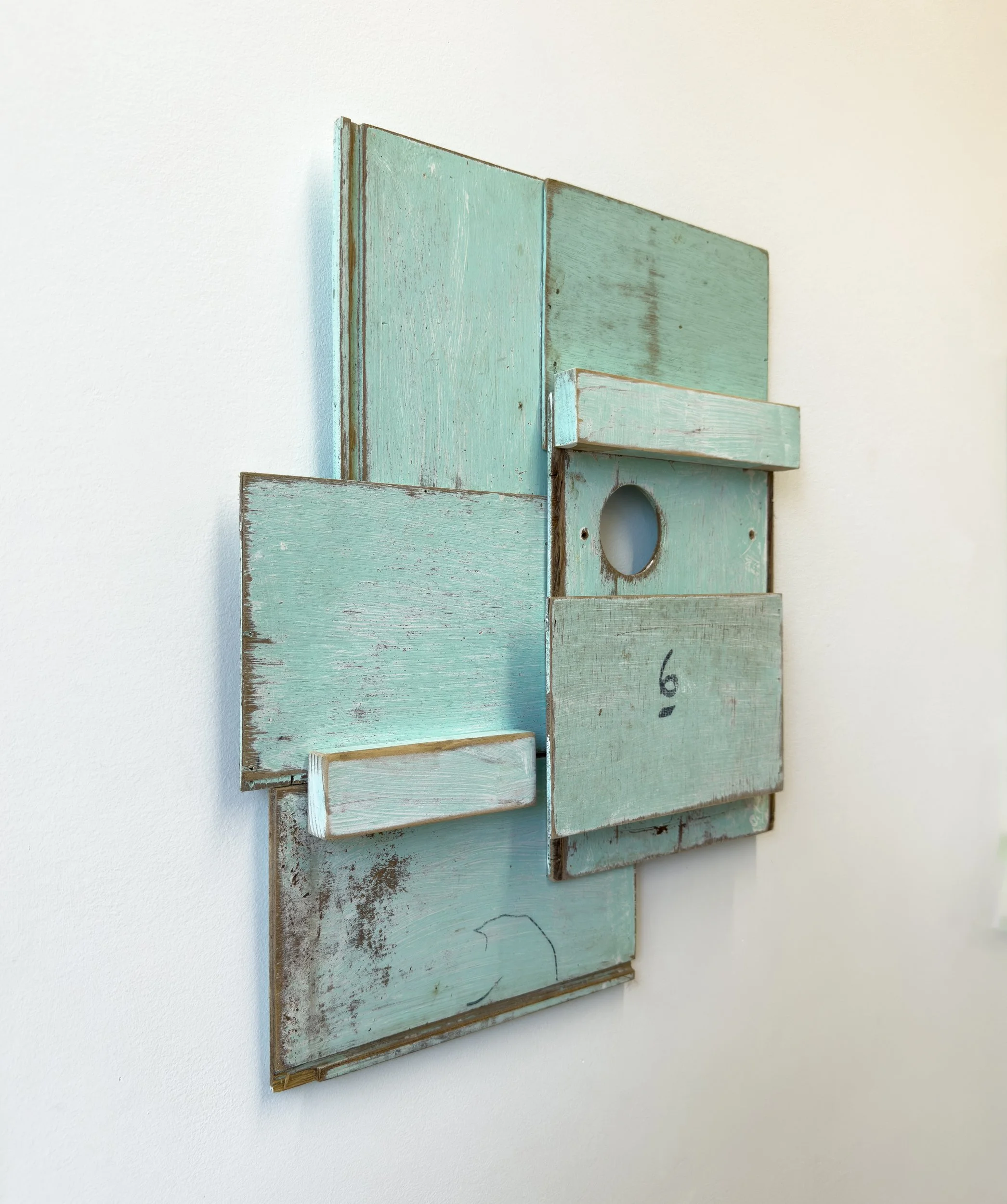 Abstract artwork composed of distressed, turquoise wooden panels arranged in geometric shapes. Features a circular cutout, creating a rustic feel.