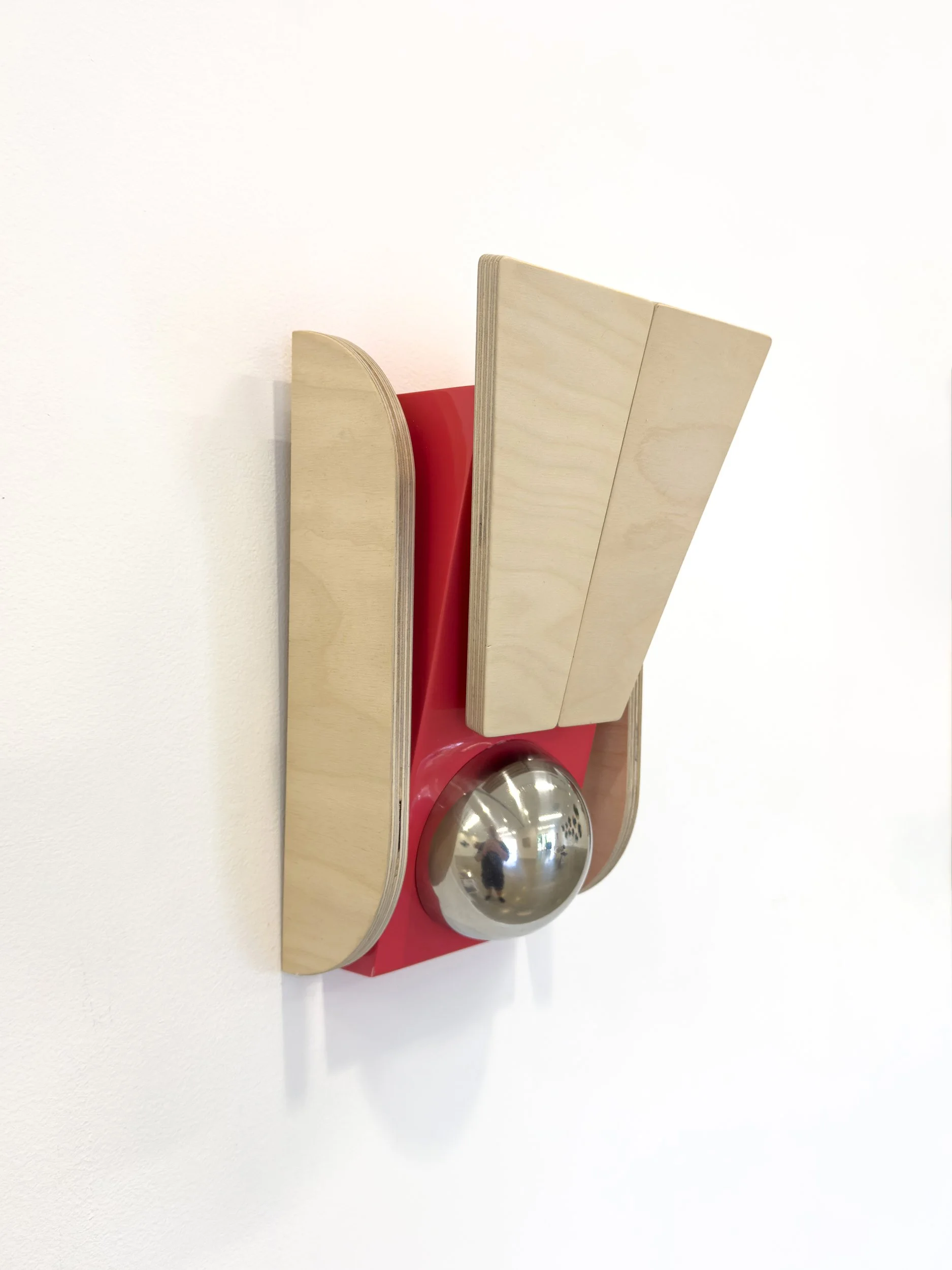 Wall-mounted abstract sculpture with beige wood shapes and a reflective metallic sphere on a red backing, creating a modern and geometric feel.
