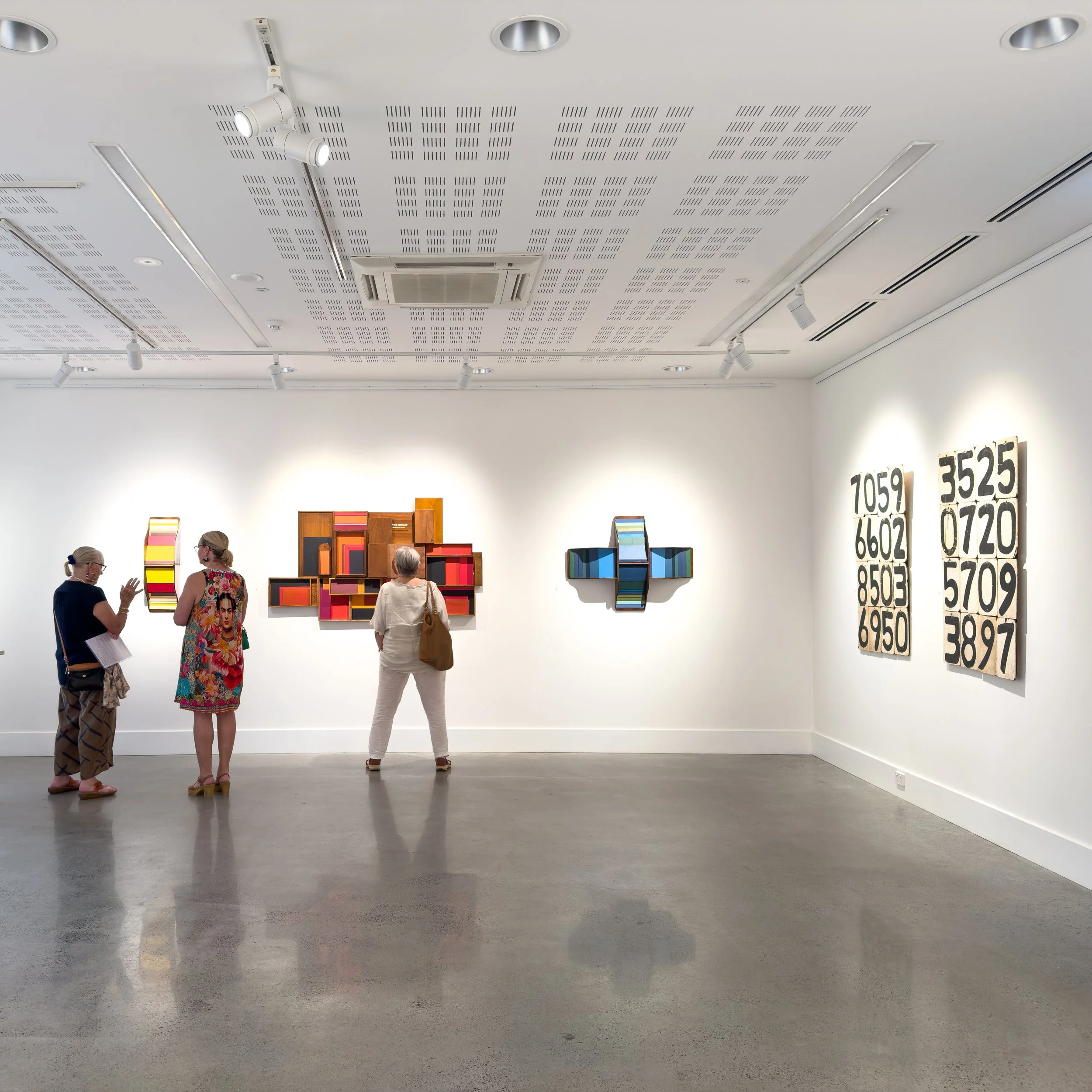 Three people in a modern art gallery observe colorful, geometric paintings on white walls. The bright, spacious room has a reflective concrete floor.