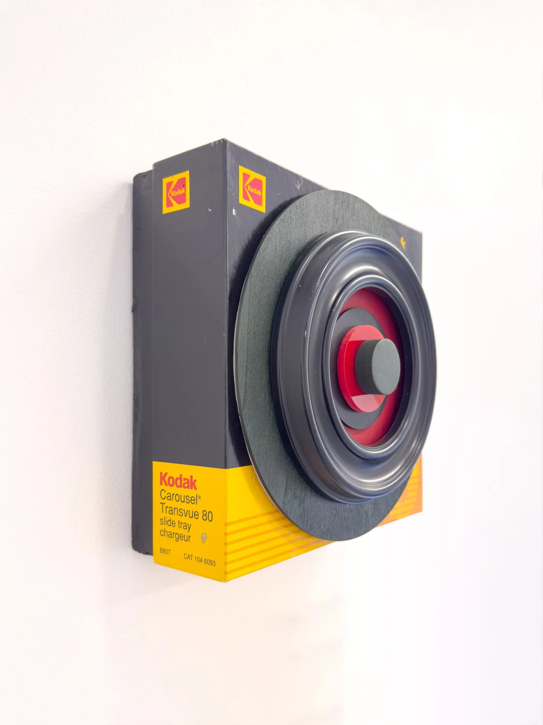 A Kodak Carousel slide tray, mounted on a white wall, features bold yellow and black packaging with red accents, evoking nostalgia and vintage vibes.