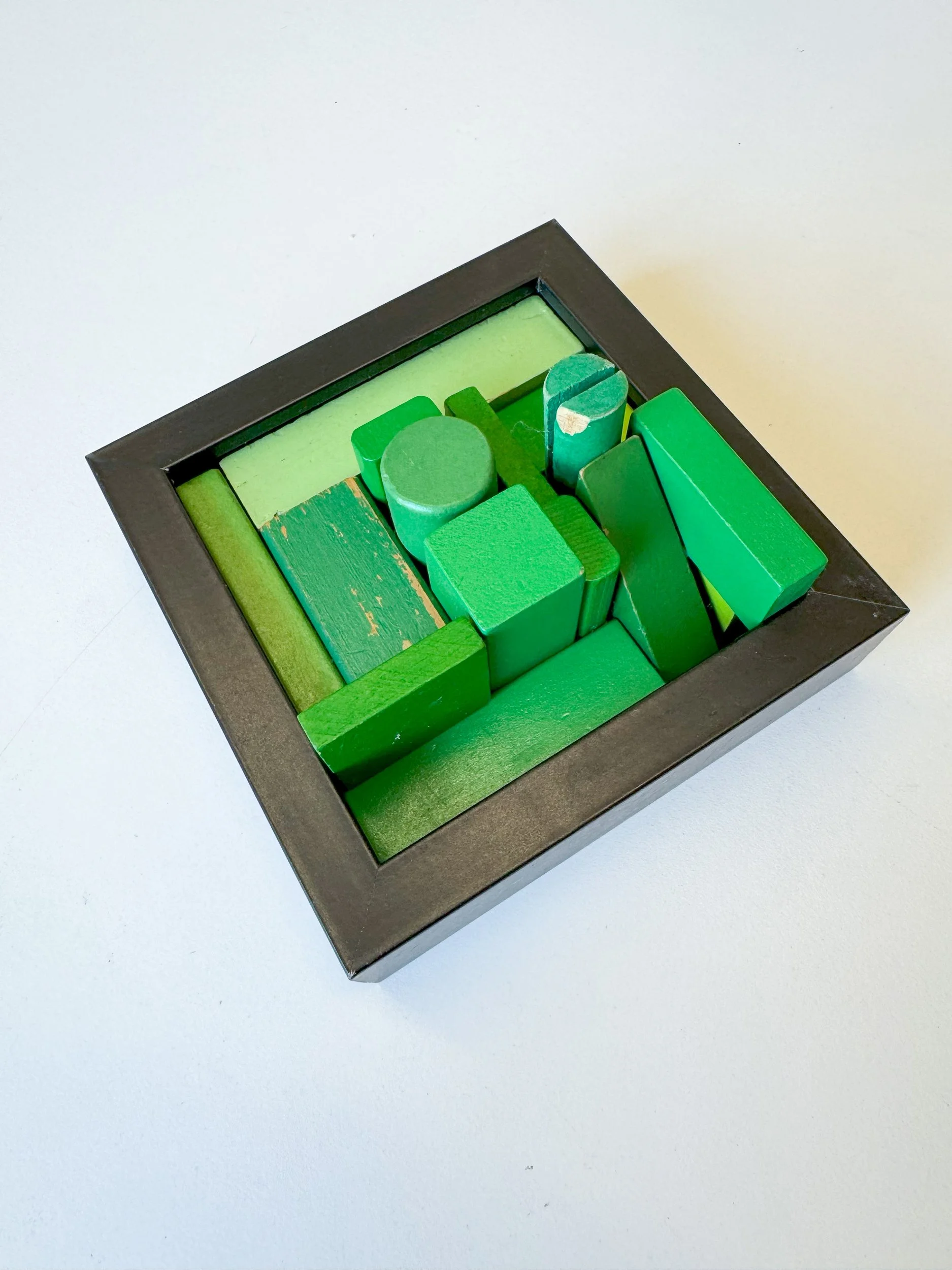 A framed artwork features assorted green wooden blocks arranged in a geometric pattern. The varying shades and shapes create a modern, abstract composition.