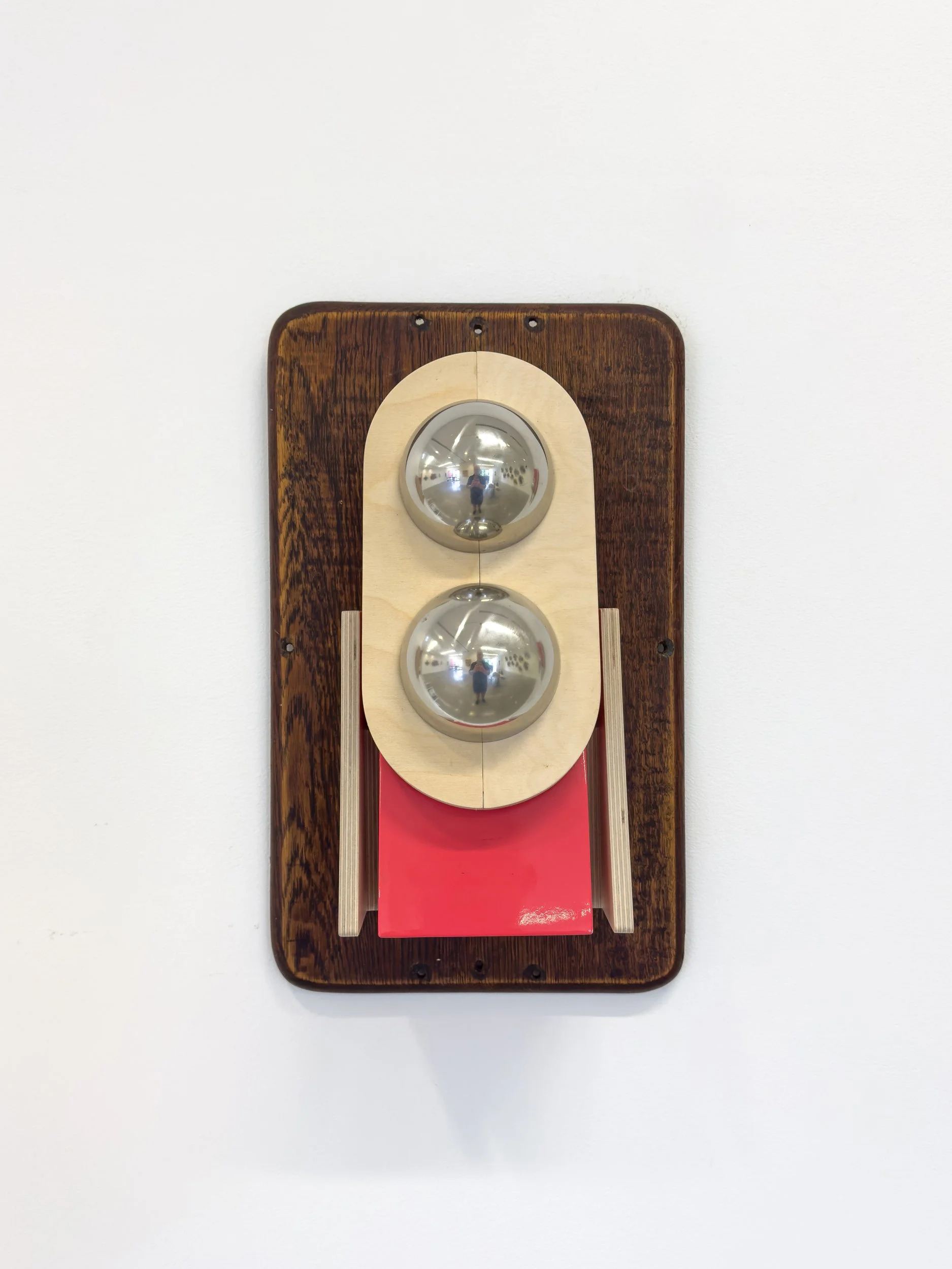 Wooden wall-mounted sculpture with two metallic spheres in the center. A red panel rests beneath in a minimalist design against a white background.