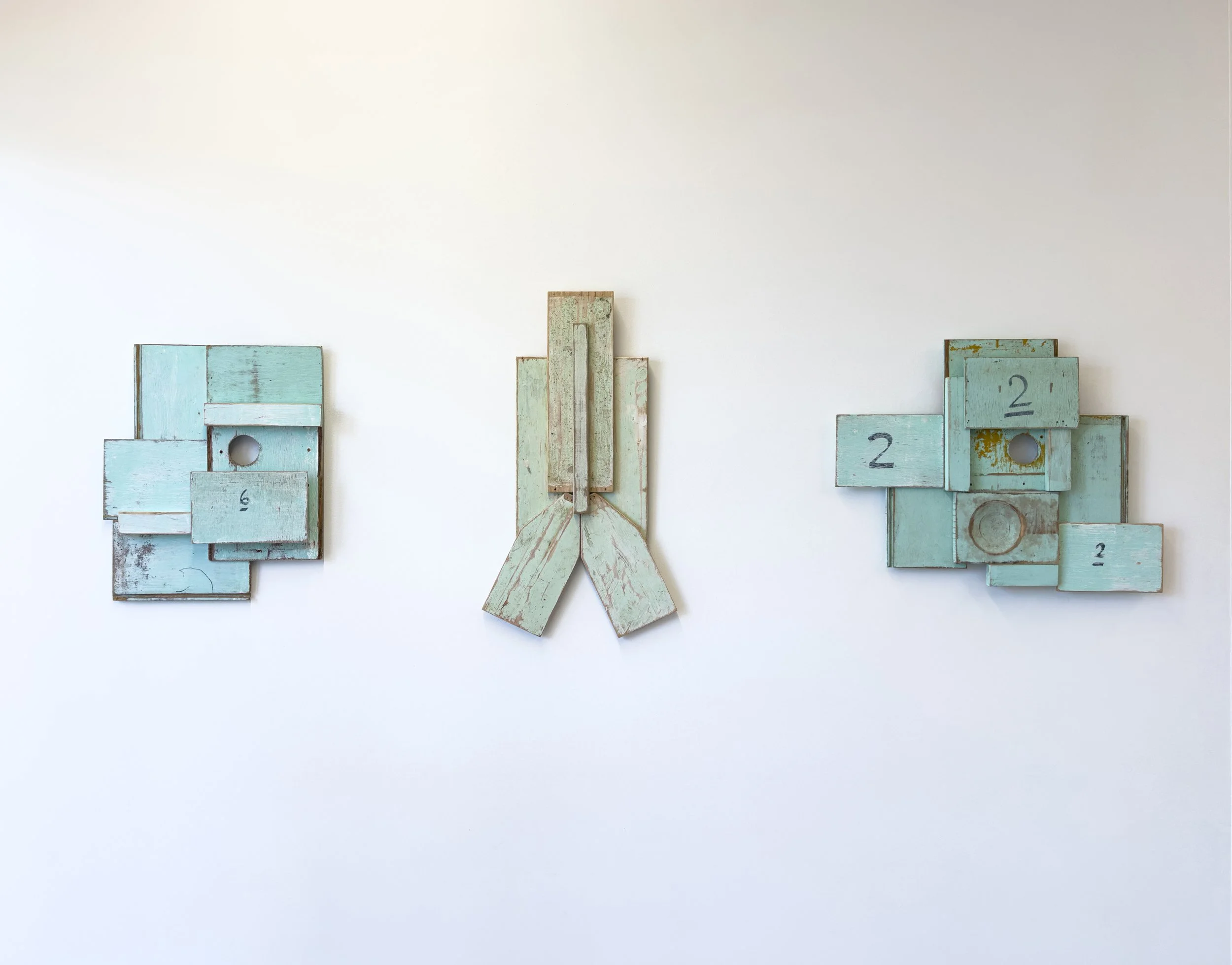 Three abstract artworks with distressed teal wood pieces form geometric shapes on a white wall. Each piece has numbered elements, conveying a rustic, artistic tone.
