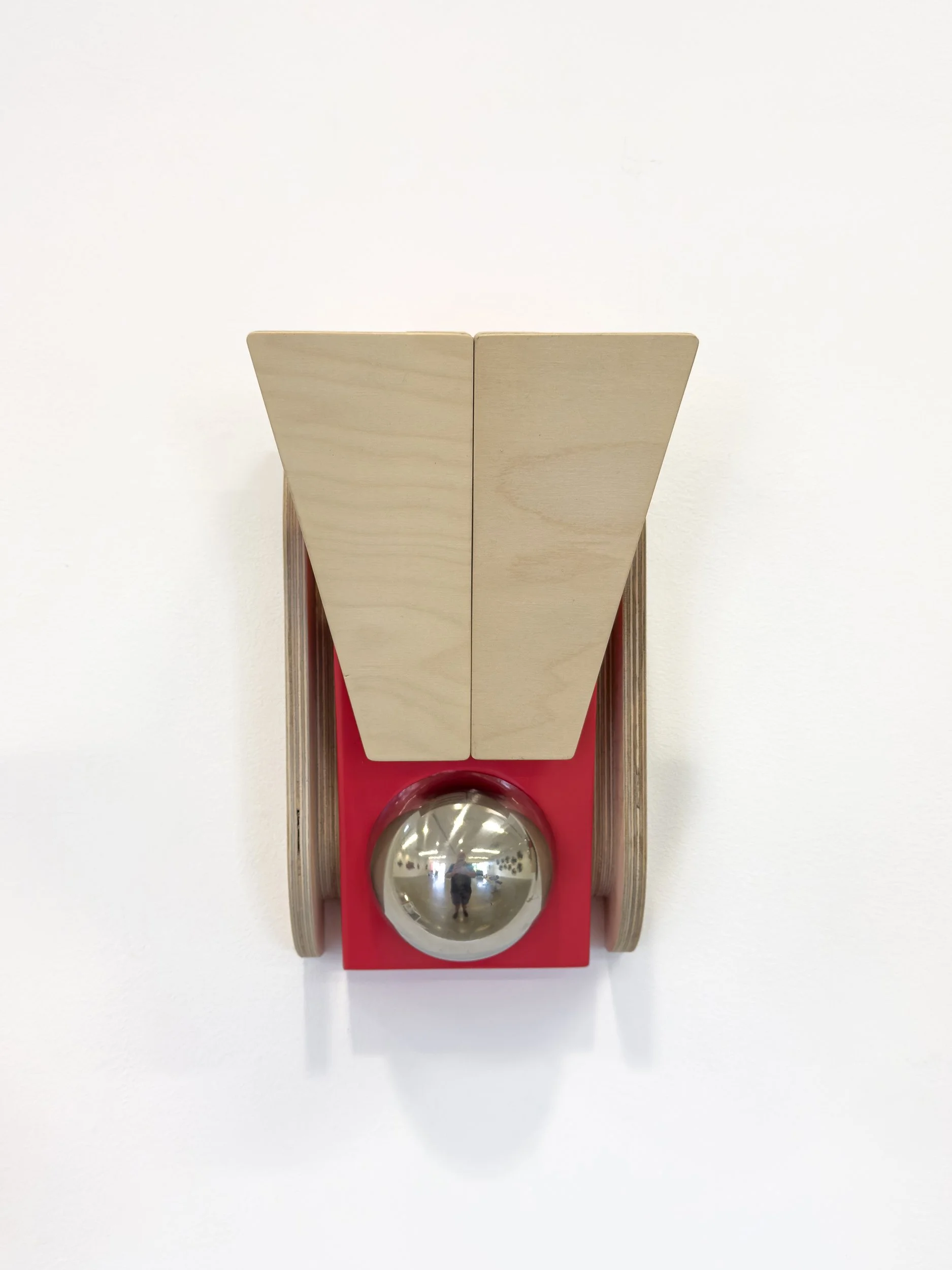 Modern sculpture on a white wall featuring a red base with a metallic sphere and two vertical wooden panels, creating a sleek and minimalist design.