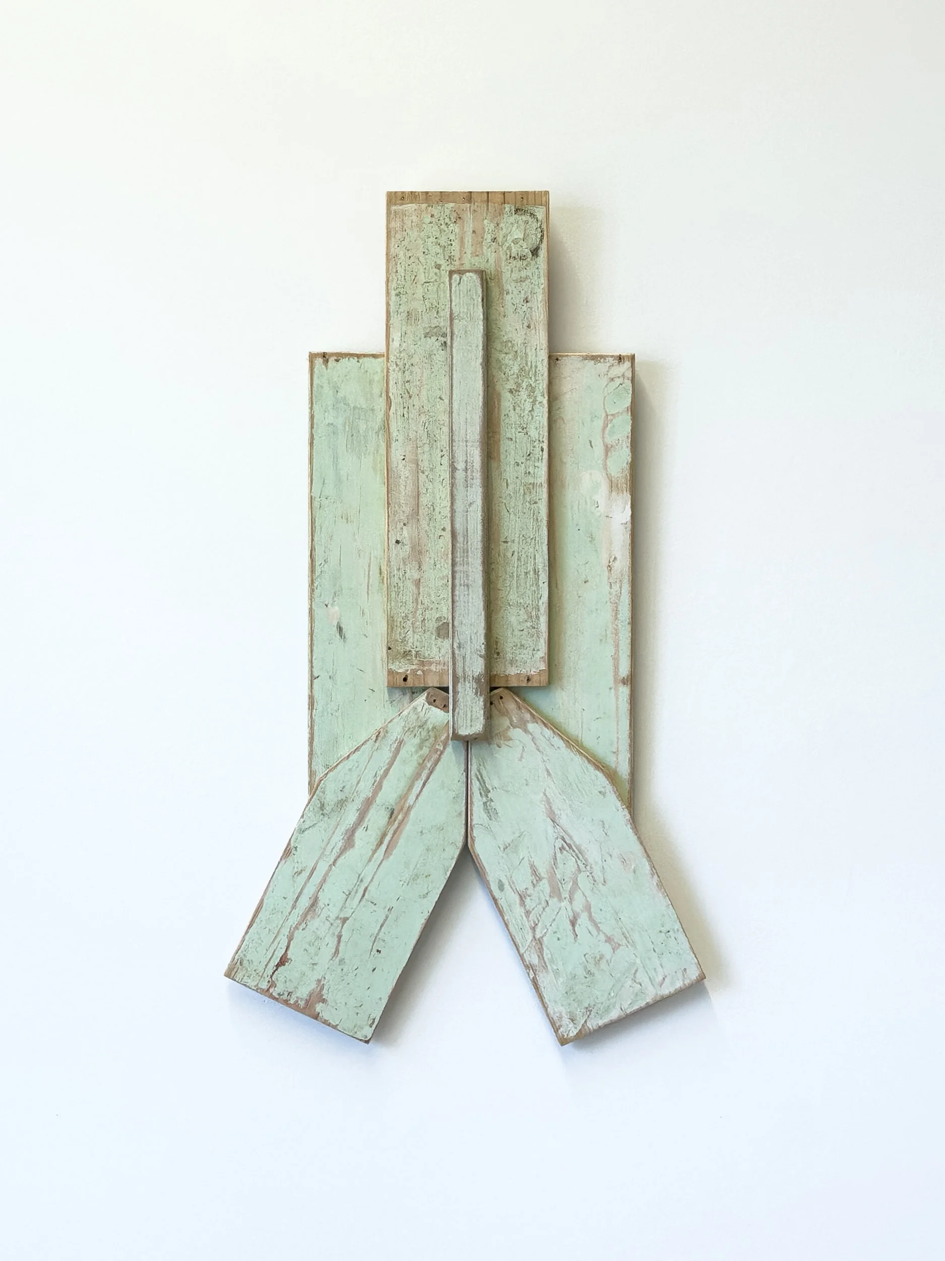 Abstract sculpture of overlapping, weathered wooden planks in green and brown, forming a geometric shape against a white background. Minimalist and rustic.