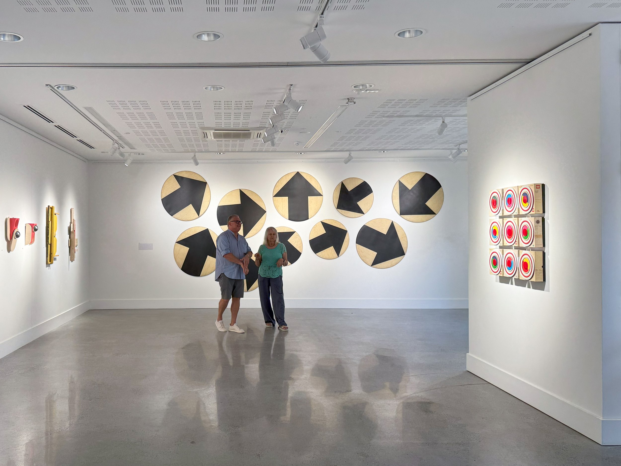 Two people stand in a bright art gallery with geometric black and yellow circular artworks on the wall and a set of pink target-like patterns on the right.