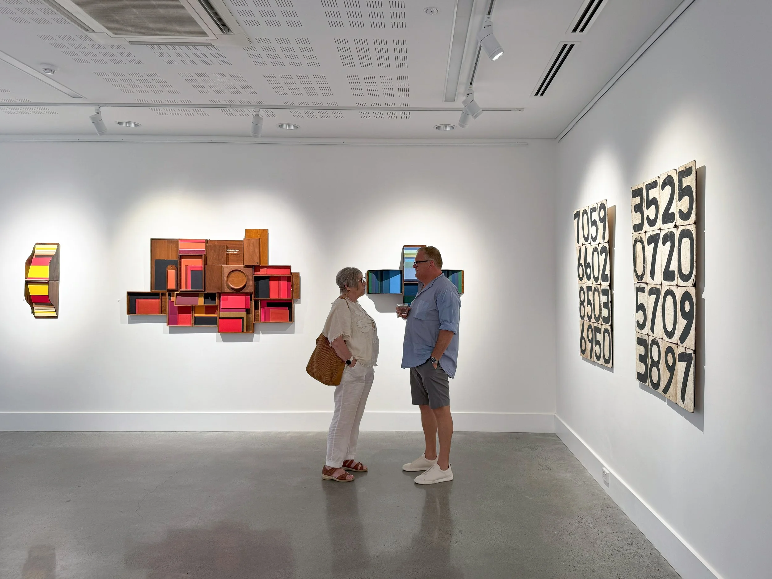 A woman and man converse in an art gallery. They stand in front of abstract artworks with vibrant colors and numbers, creating a contemplative atmosphere.