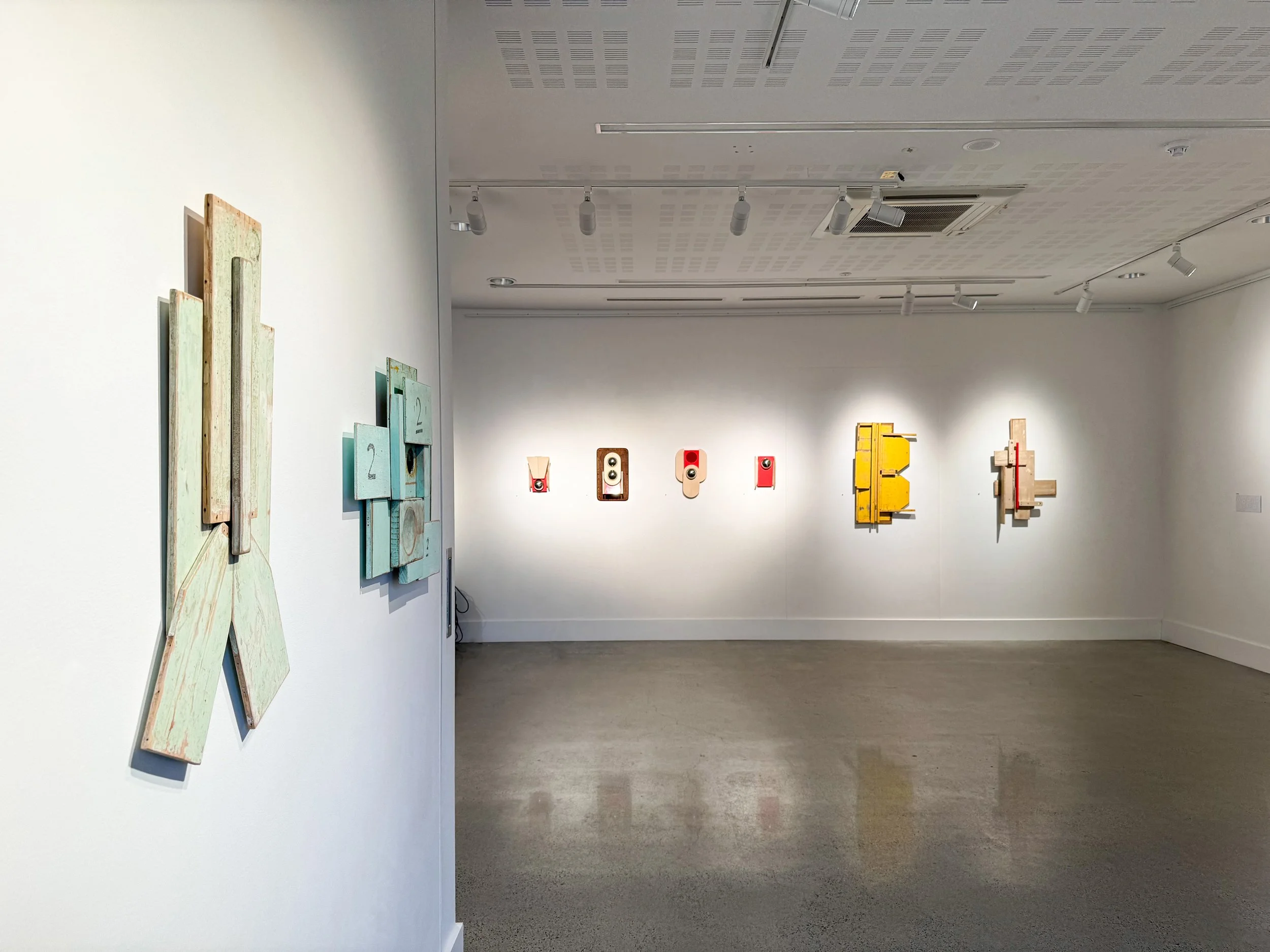 A modern art gallery with geometric wood sculptures on white walls. The pieces vary in color, including teal, red, and yellow. The room is well-lit and spacious.