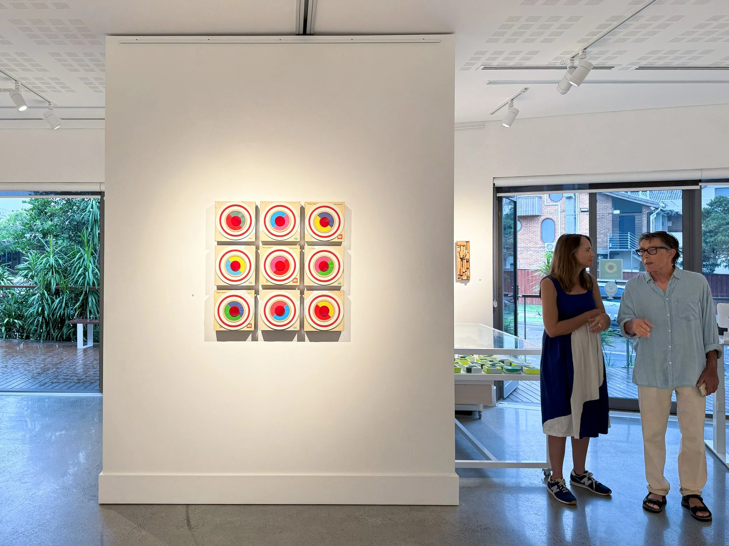 Gallery exhibition of circular assemblages with two visitors viewing the work under soft lighting