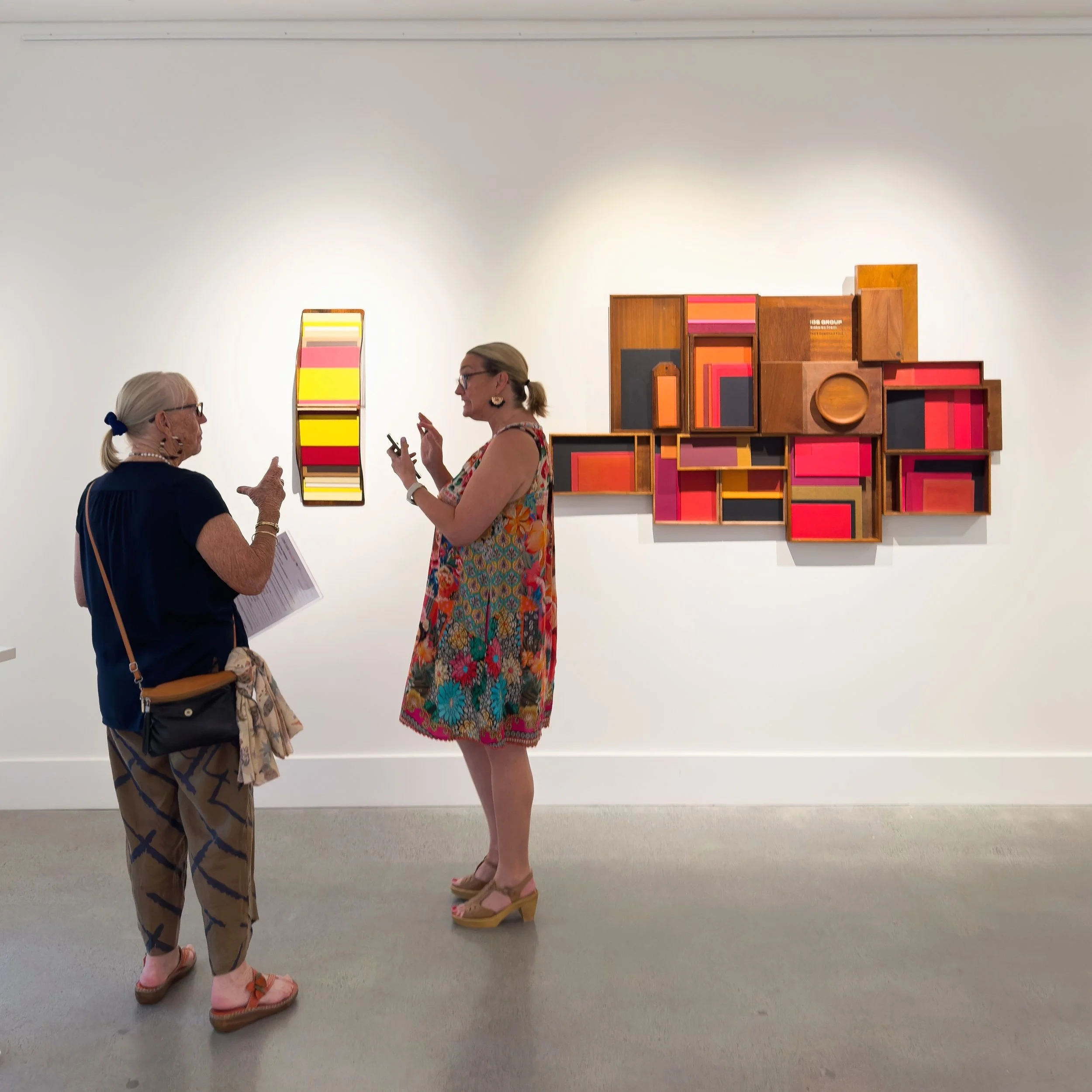Two women engaged in conversation in an art gallery. They're viewing colorful, geometric wall sculptures in red, yellow, and brown tones. The atmosphere is lively and thoughtful.