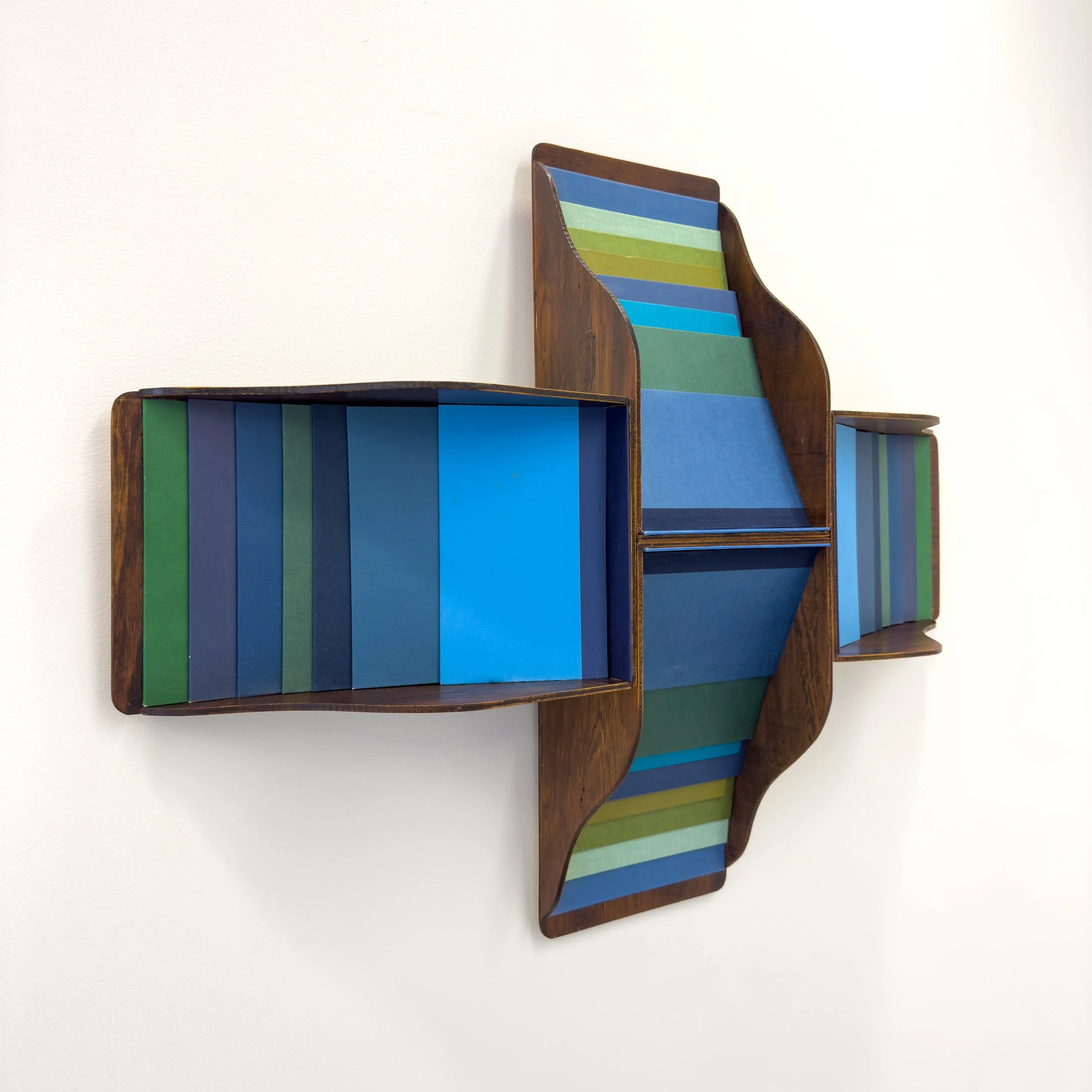 Wooden wall-mounted shelf with wavy edges, holding colorful panels in shades of blue and green. Modern aesthetic on a white background.