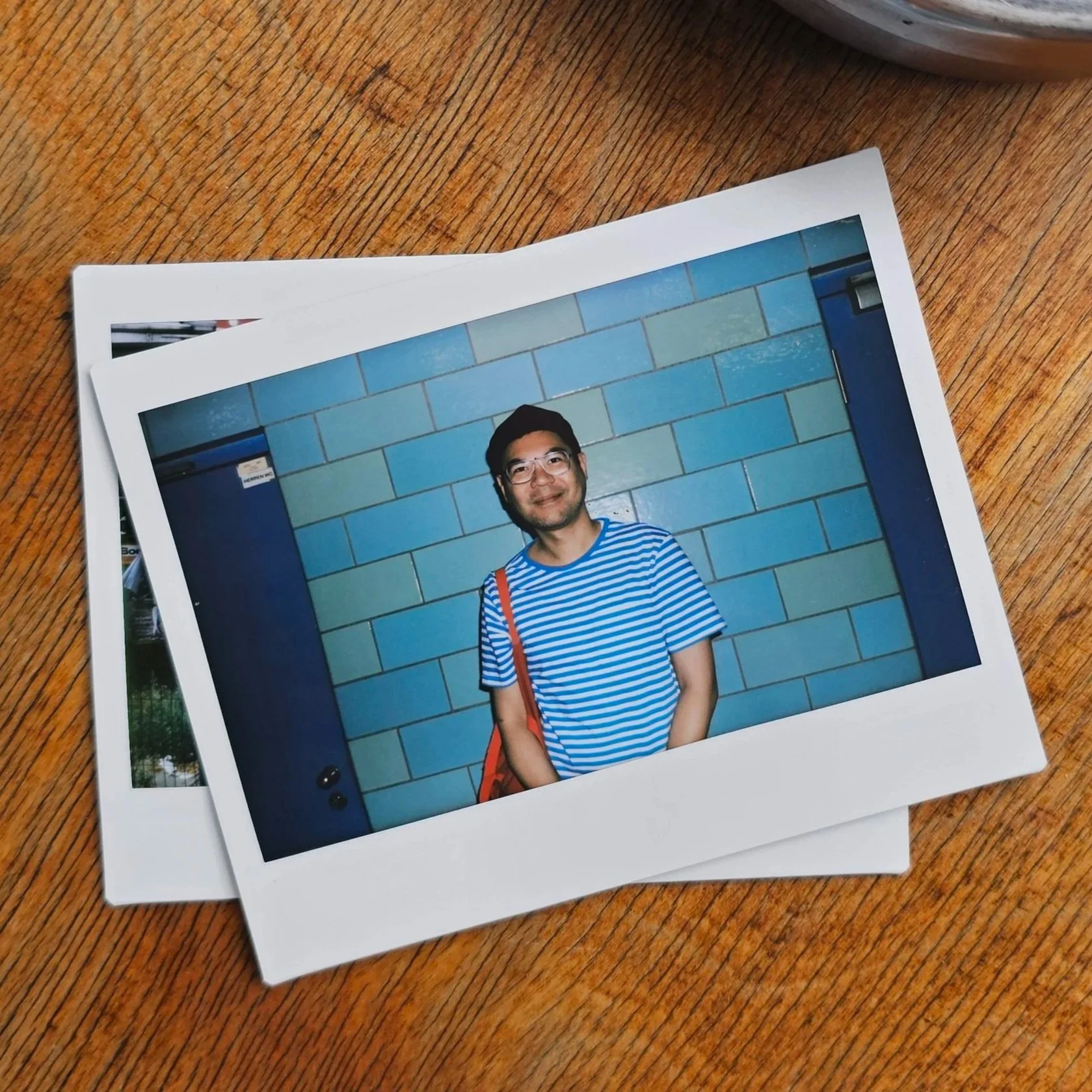 A small pile of polaroid photographs on a wooden table. The first depicts me, the anthropologist, professor, and creative director Christopher Santo Domingo Chan, standing before the blue tiles of the Alexanderplatz U-bahn station in Berlin, Germany.