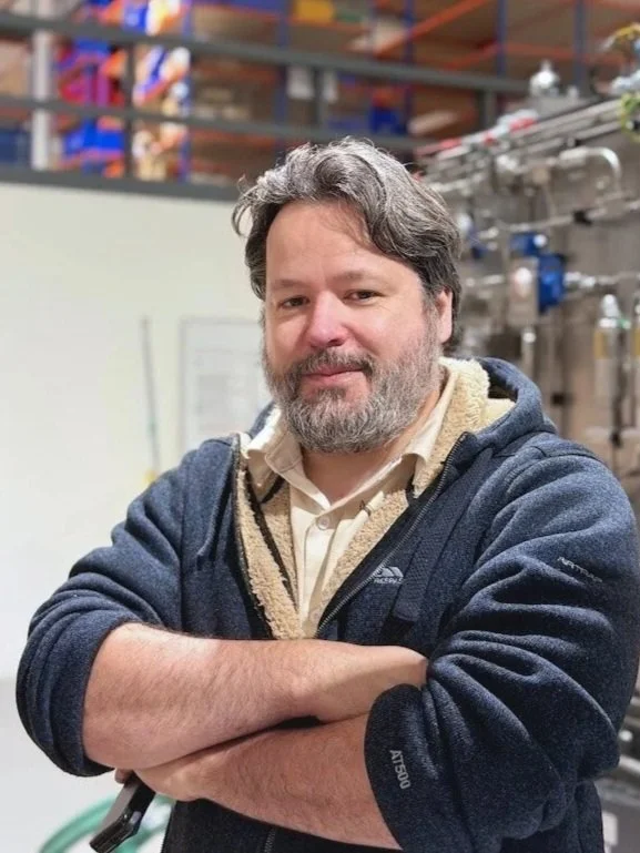 Person standing in front of a brick wall wearing a black jacket and blue shirt, with a logo on the jacket.