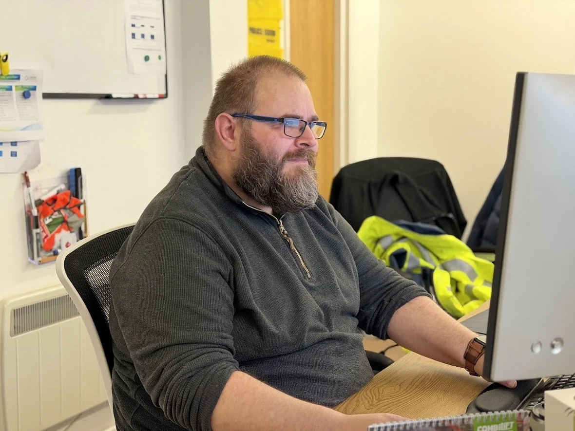 A man with glasses and facial hair working on a computer in an office setting, wearing a blue shirt.