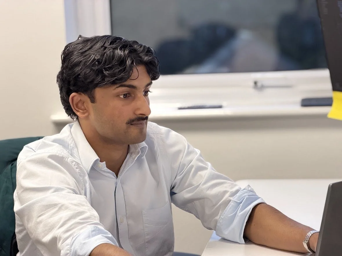 Man in white shirt with dark hair, standing indoors with plants in the background.
