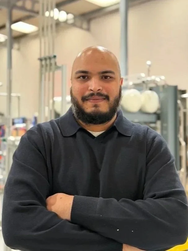 Person wearing a black jacket with "Fuel Cell Systems" logo, standing in front of a red brick wall.
