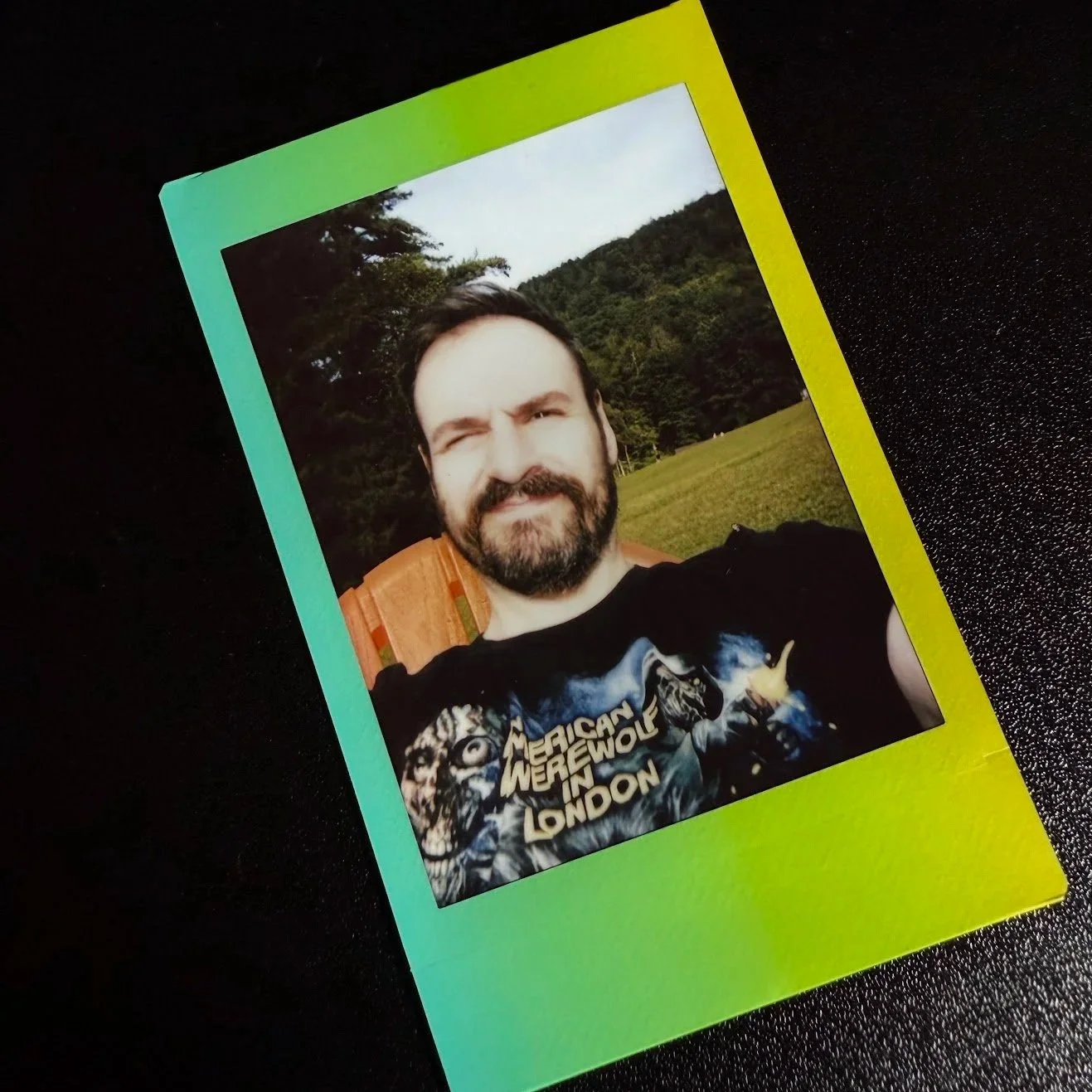 Polaroid photo of a man with a beard outdoors, wincing or squinting, wearing a black T-shirt with a graphic design that says 'Mexican Werewolf in London', with trees and a hill in the background.