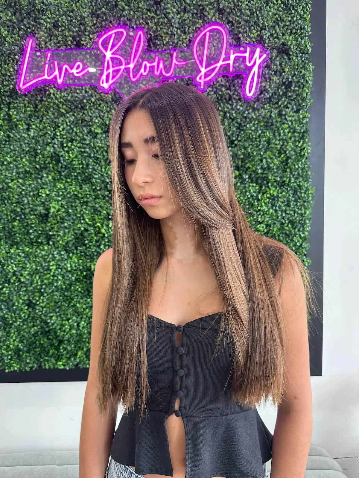 Client with a smooth brunette blowout in front of the Live Blow Dry neon sign