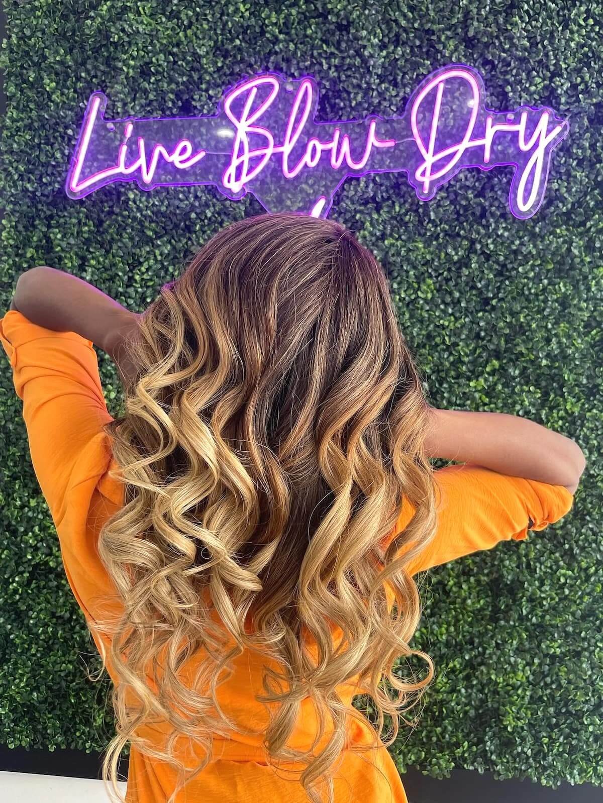 Curled blonde blowout in front of the pink Live Blow Dry neon sign