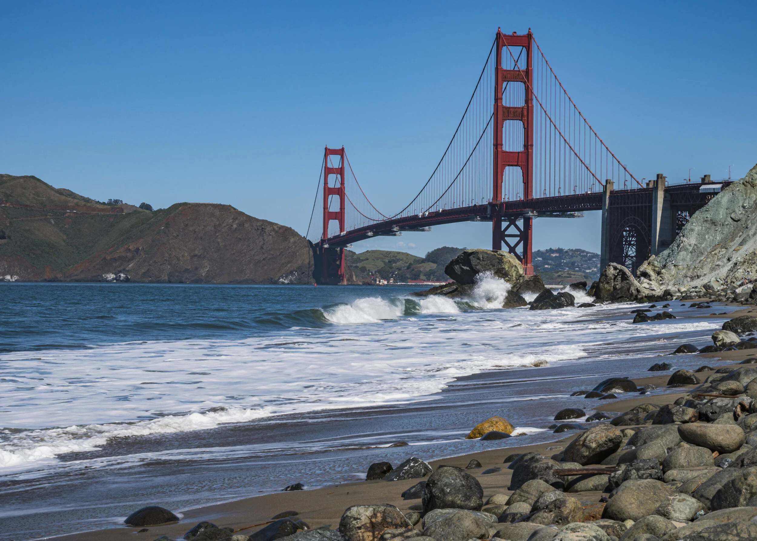 View of the Golden Gate Bridge over the water with rocky beach in the foreground and hills in the background on a clear day.