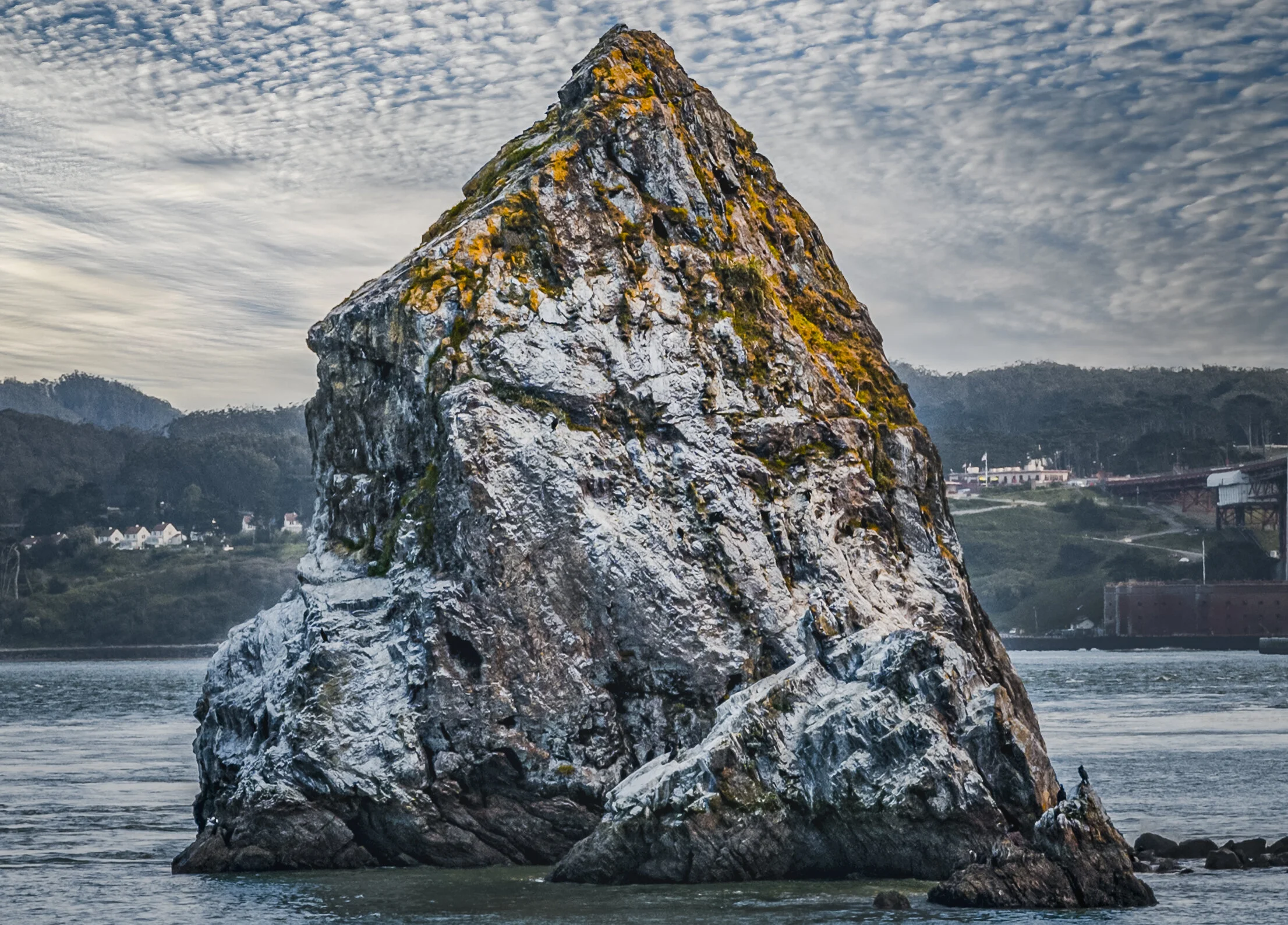 Large rocky sea stack with moss and lichen, tallest point peaks upward, surrounding water and distant hilly shoreline with houses, under a cloudy sky.