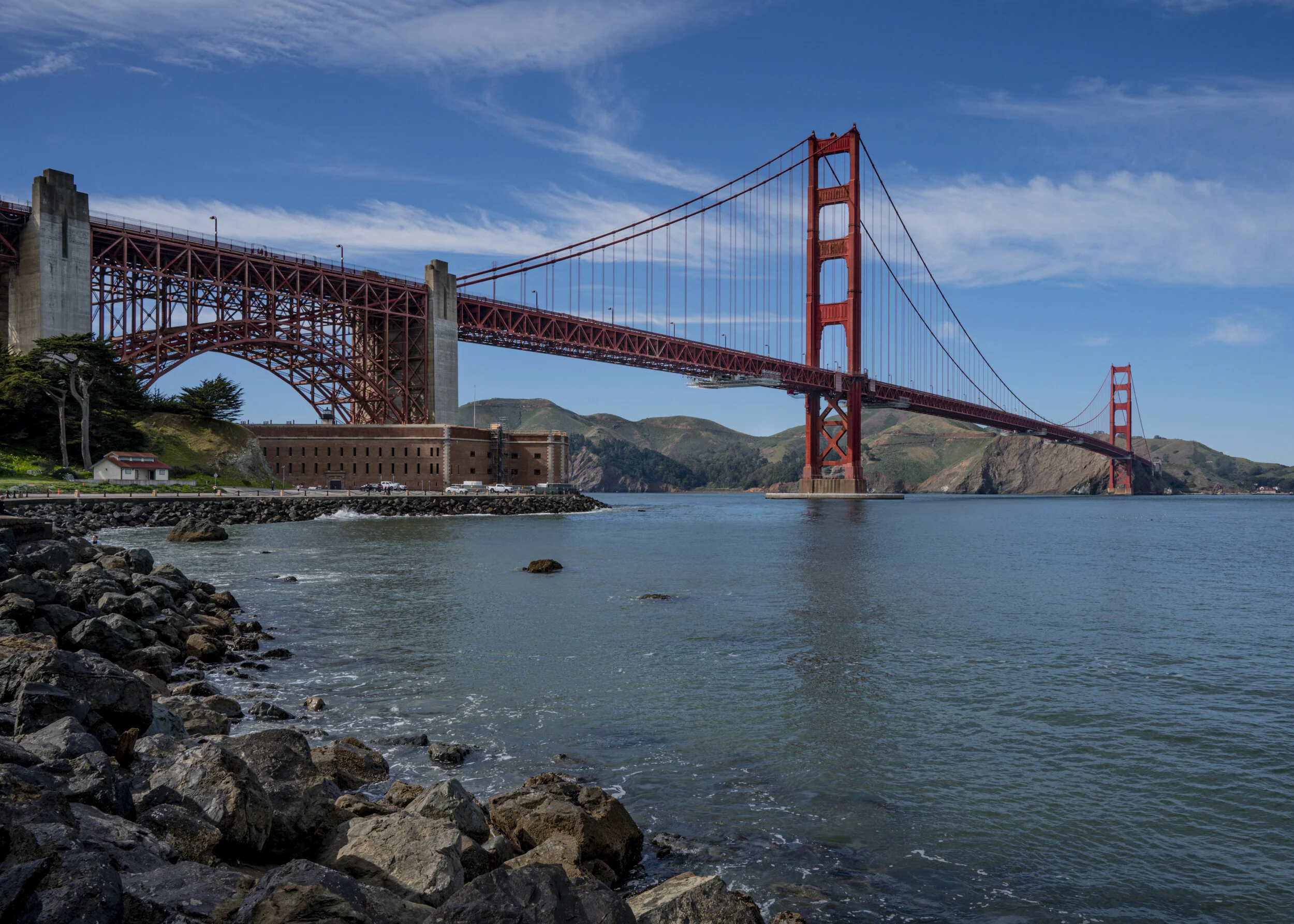 Photograph of the Golden Gate Bridge in San Francisco, California, spanning the water with hills in the background and rocks in the foreground.