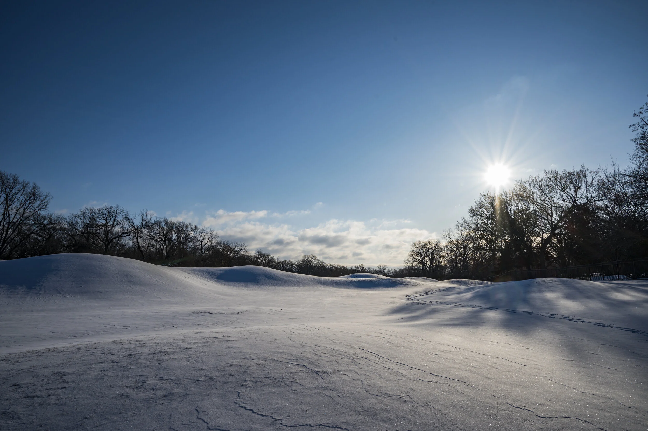 Snow-covered landscape with small hills under a blue sky with scattered clouds, and the sun shining brightly near the horizon.