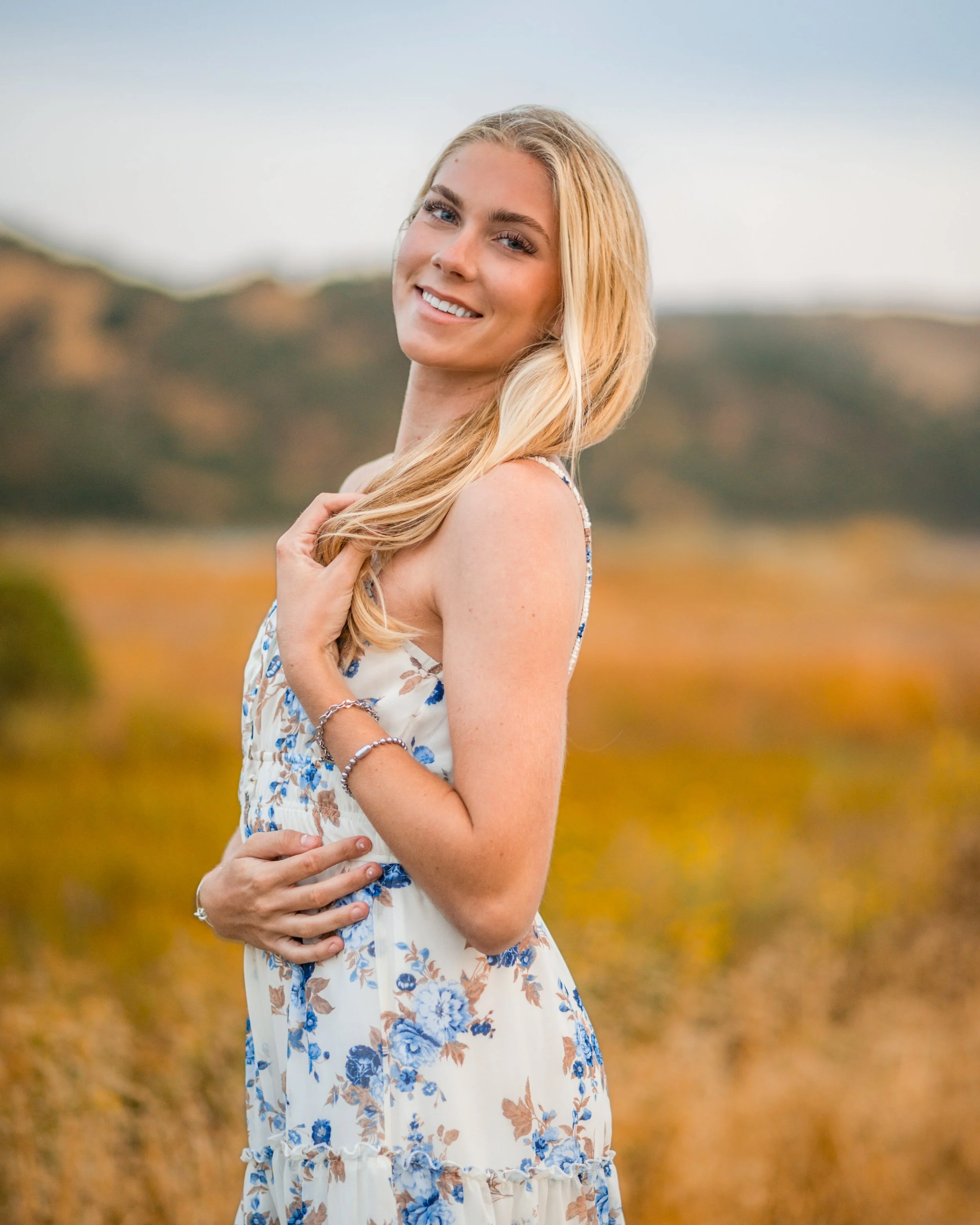 A young woman with long blonde hair smiling outdoors in a field with hills in the background, wearing a sleeveless floral dress.