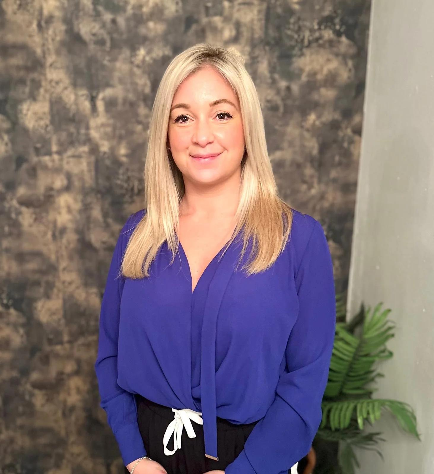 A woman with long blonde hair wearing a royal blue blouse and black pants with a white drawstring tied at the waist, standing in front of a textured dark wall and green plants.