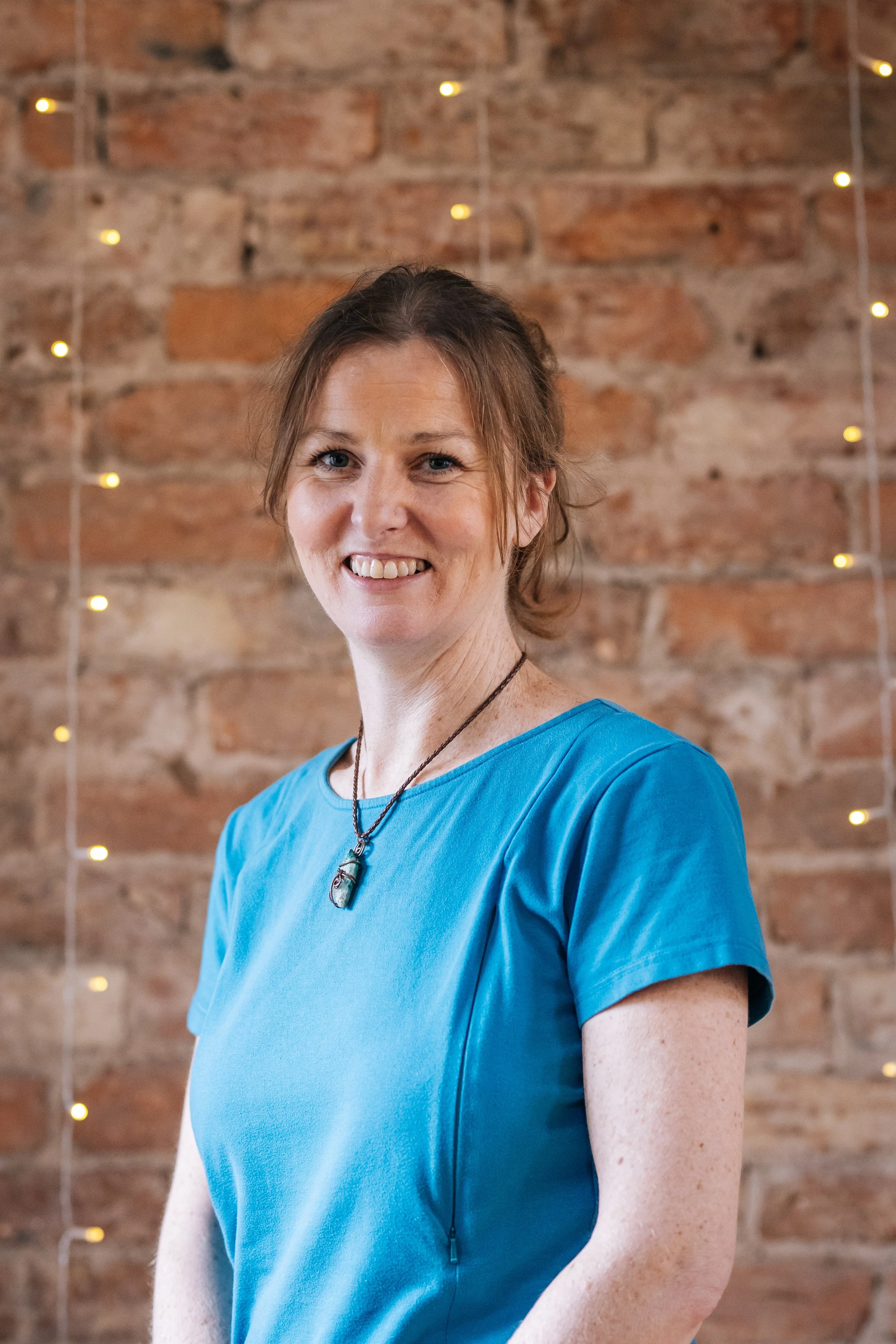 A smiling woman with brown hair in a blue shirt stands in front of a brick wall decorated with string lights.