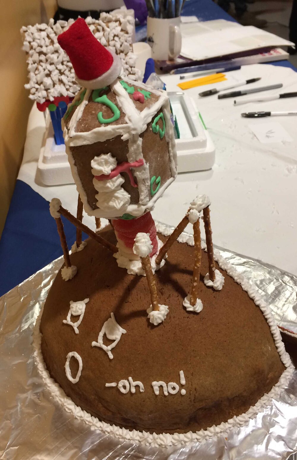  The back of the domed gingerbread “cell” with geometric “phage” on top - drawn in white frosting are two eyes and a round mouth, saying “oh no!” 